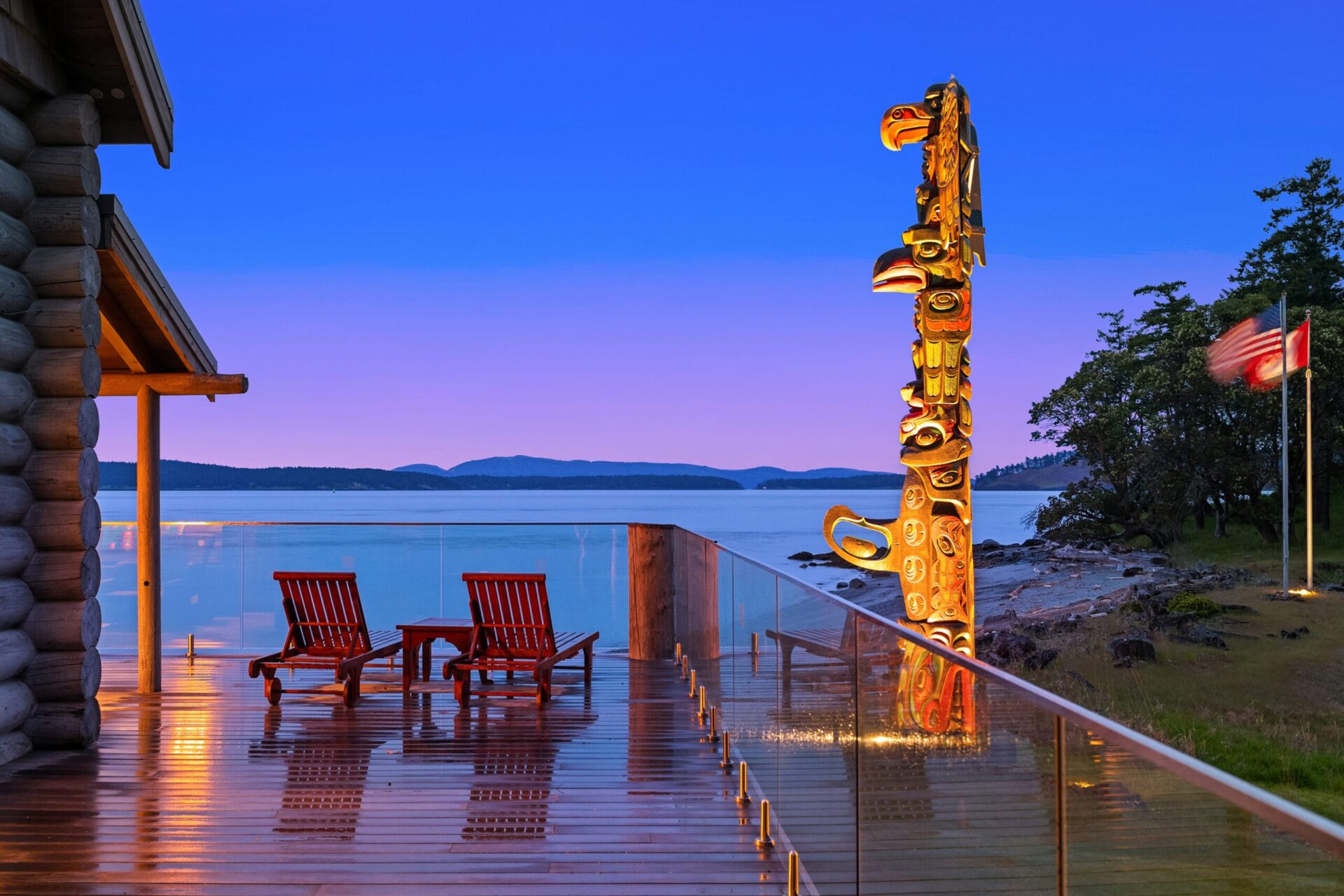 A serene lakeside scene at sunset features an hdr wooden deck with two empty lounge chairs, a totem pole, and a reflecting glass railing. The calm lake and distant mountains enhance the peaceful atmosphere, with a wooden cabin partially visible on the left. Perfect for top real estate listings. Twilight replacement.