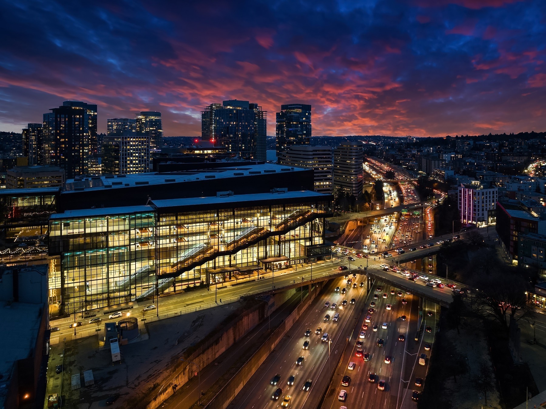 A vibrant cityscape at dusk featuring brightly lit office buildings and a busy multi-lane highway filled with traffic. The sky is ablaze with hues of orange, pink, and purple as the city lights begin to illuminate the night.