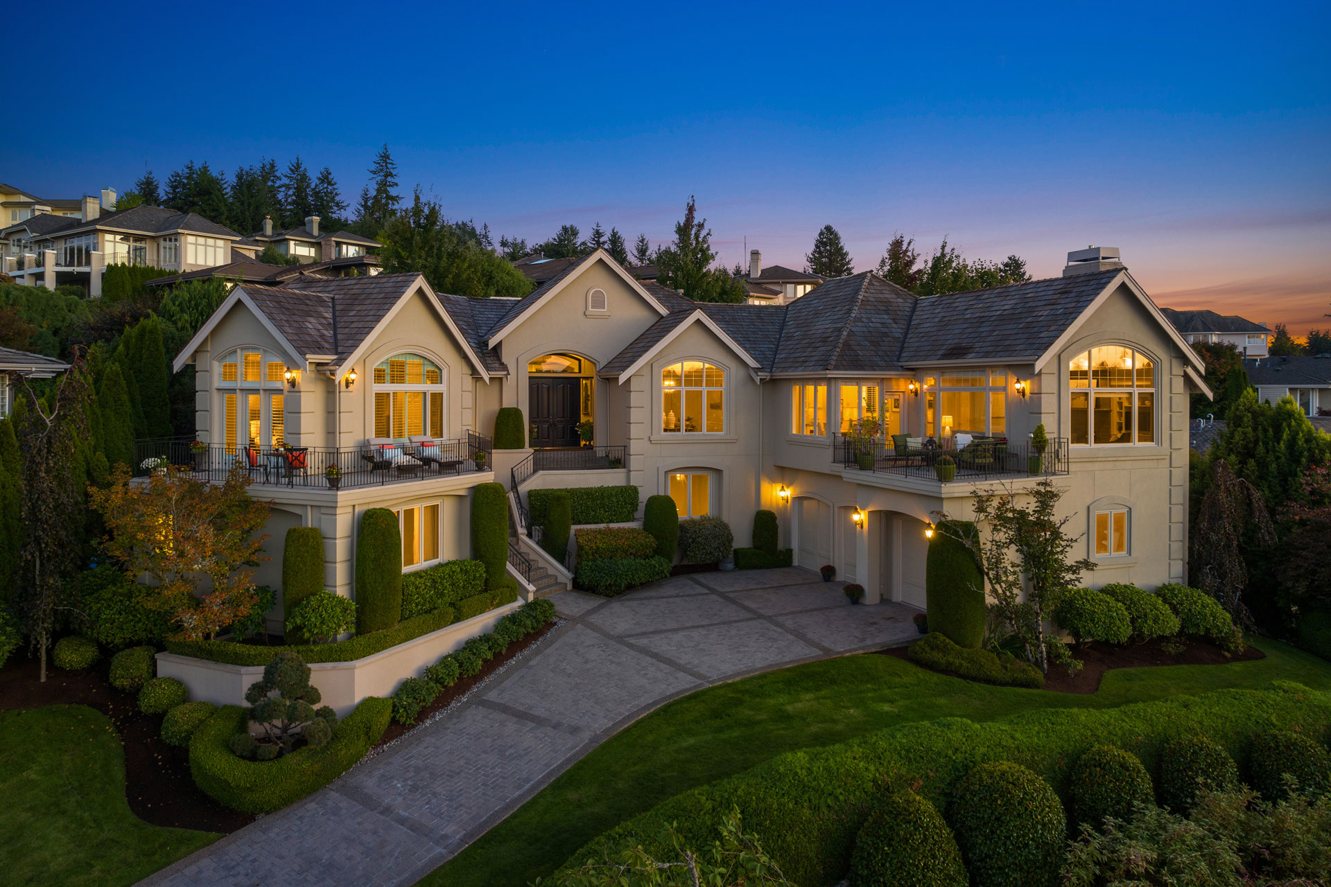 A grand two-story house illuminated at dusk, featuring a spacious driveway, well-manicured landscaping, and large arched windows. The home includes multiple balconies and is surrounded by greenery, with neighboring houses visible in the background.