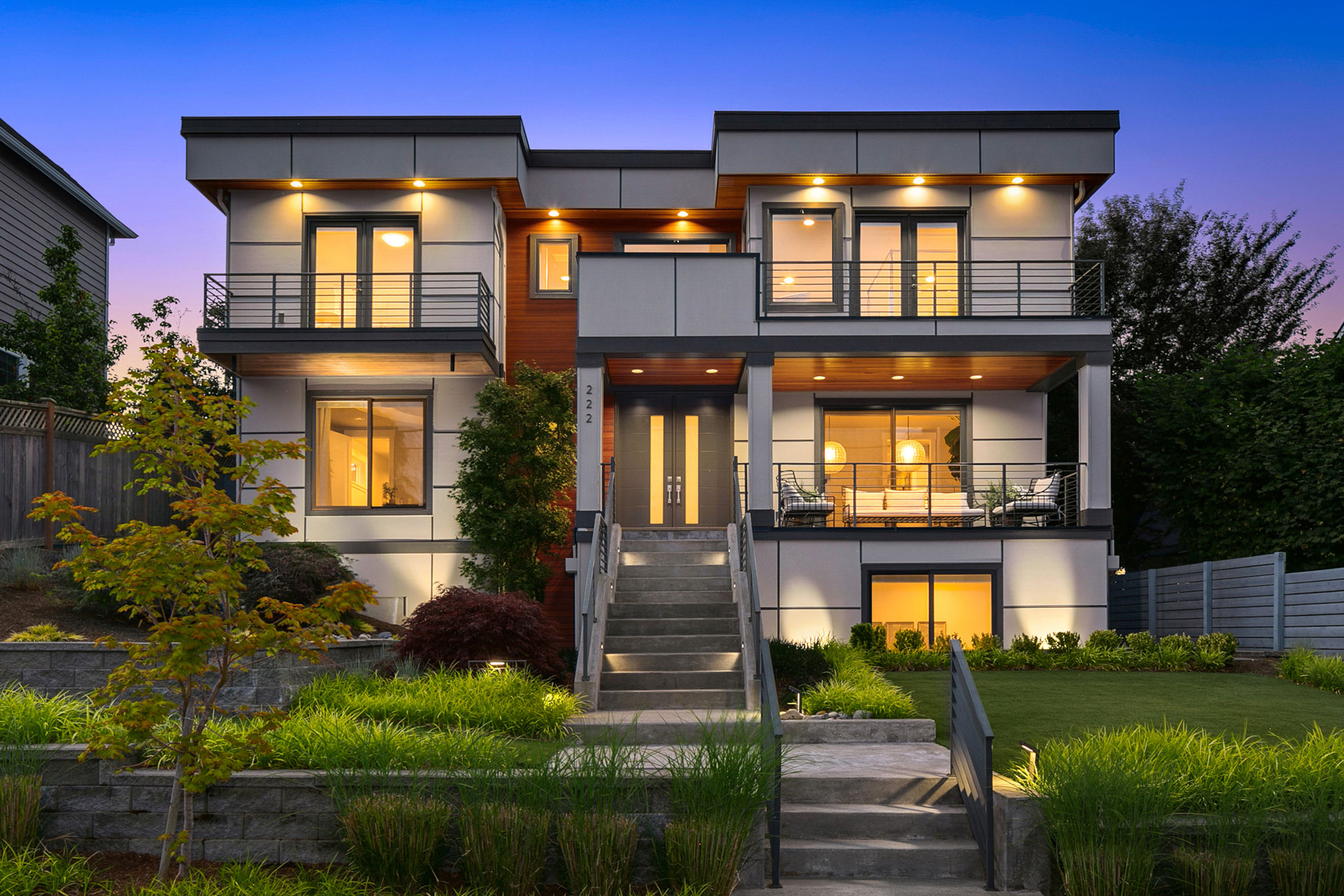 A modern two-story house with a flat roof and an array of windows and balconies. The exterior features a mix of white, wood, and gray panels. The front yard is landscaped with trees and bushes, and a wide staircase leads up to the front entrance. The sky is at dusk.