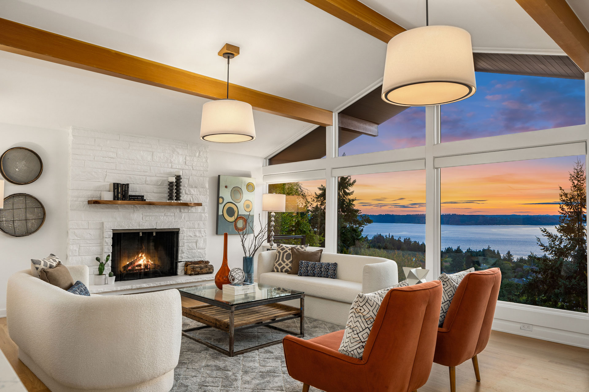 A modern living room with white walls and wooden ceiling beams featuring a large window showcasing a sunset over a coastal view. The space includes a white brick fireplace, stylish white and orange furniture, a coffee table, and contemporary decor accents.