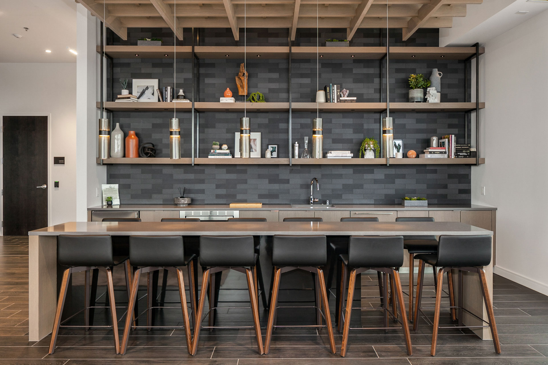 A modern kitchen with a large island counter and six bar stools. The background features open shelves with various decorative items, books, and plants. The walls are tiled with dark, rectangular tiles and the ceiling displays exposed wooden beams.