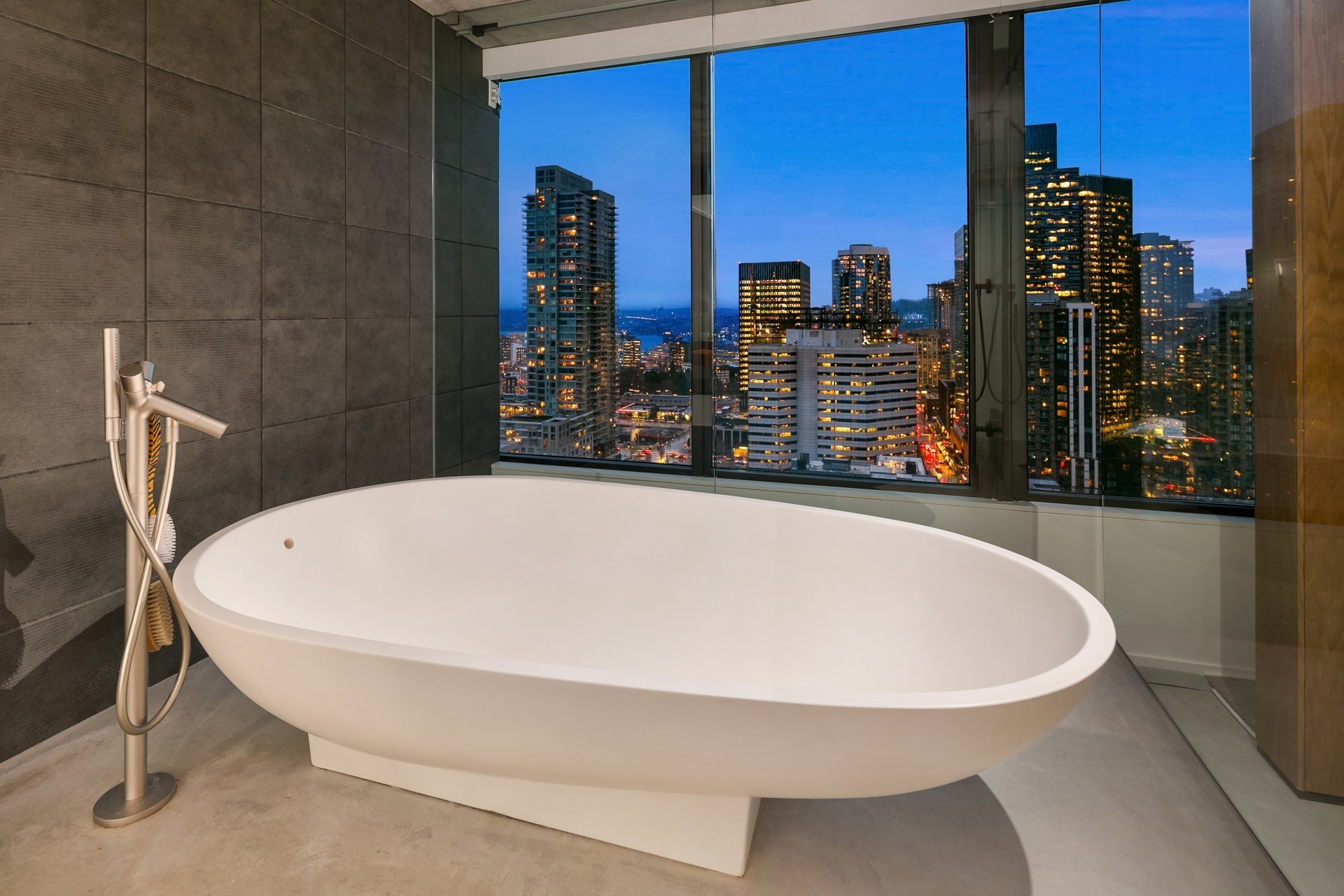 A modern bathroom with a freestanding white bathtub positioned in front of a large window offering a panoramic view of a bustling city skyline at dusk, featuring numerous tall buildings and lights. The walls are covered with dark grey tiles.