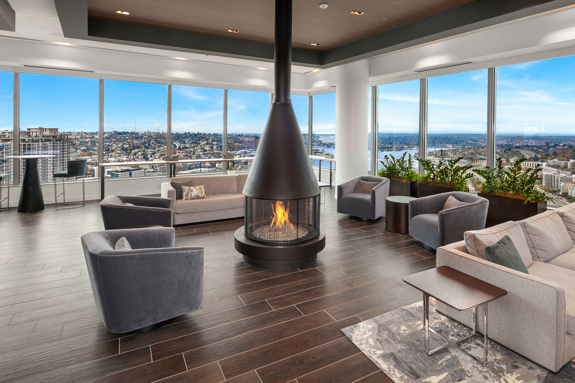 A modern living room with large windows offering a panoramic city view. The room features a central suspended fireplace, several gray armchairs, a beige sofa, and a wooden floor. Green plants near the windows add a touch of nature to the sleek design.