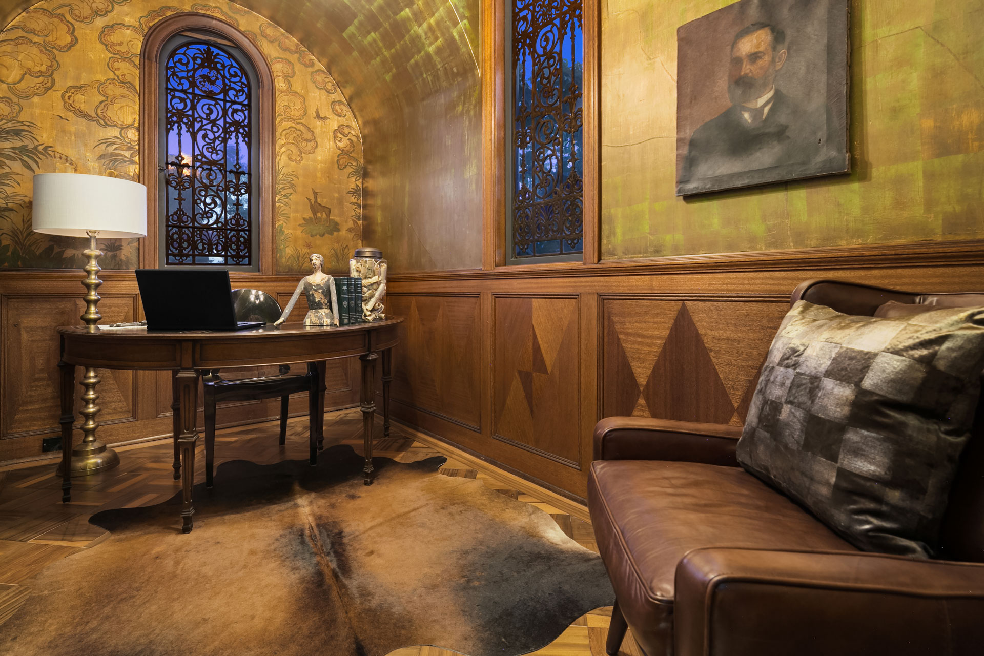 A richly decorated study room with a wooden desk, ornate lamp, and various decorative items. The walls have intricate wooden paneling and arched, stained-glass windows. A leather armchair with a patterned cushion is in the foreground, and a portrait hangs on the wall.