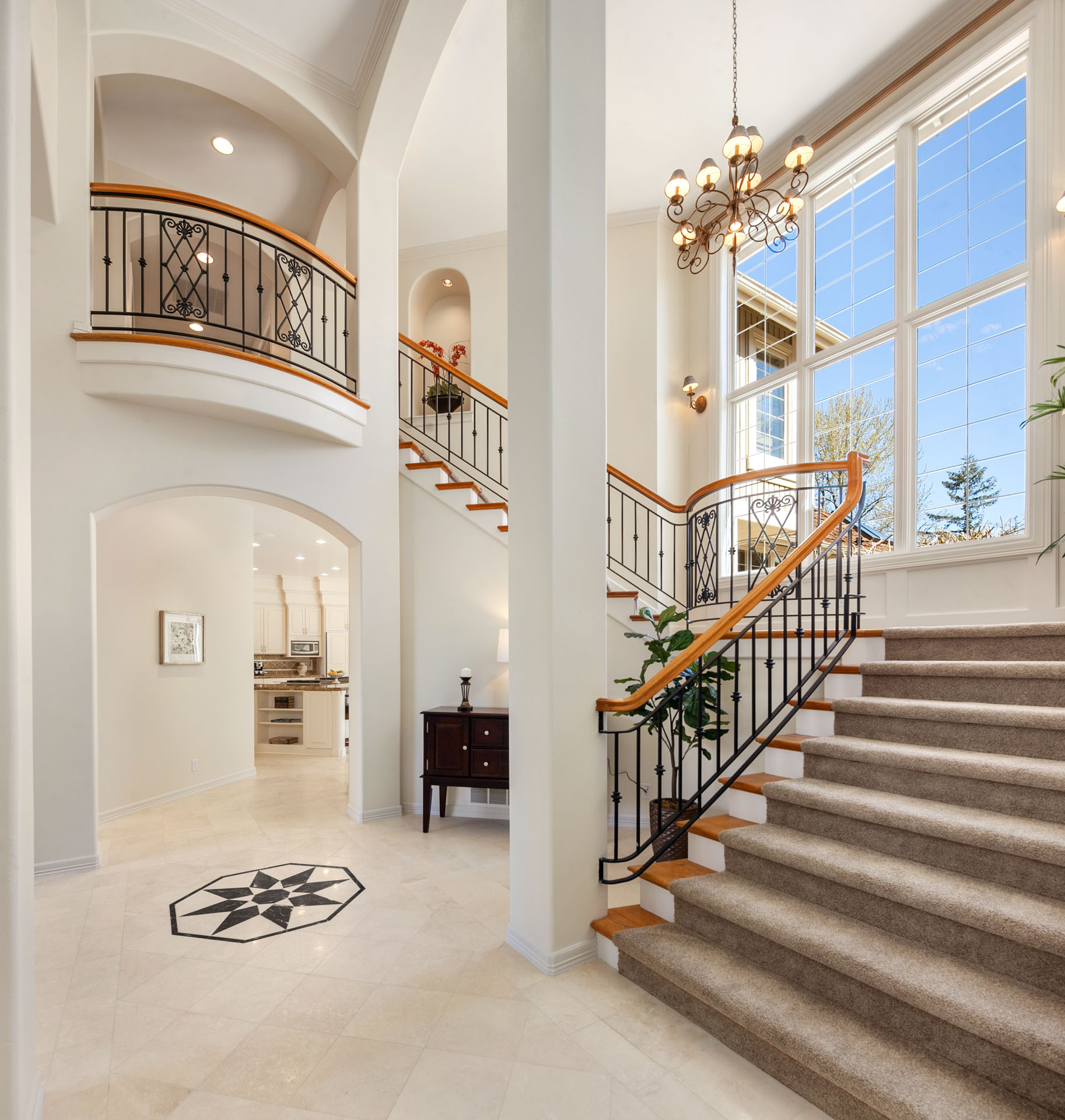 A grand, two-story foyer with a sweeping staircase featuring iron railings and wood accents. The space is bright with large windows and a chandelier hanging from the ceiling. The archway leads to the kitchen, and a dark wood console table sits against the wall.