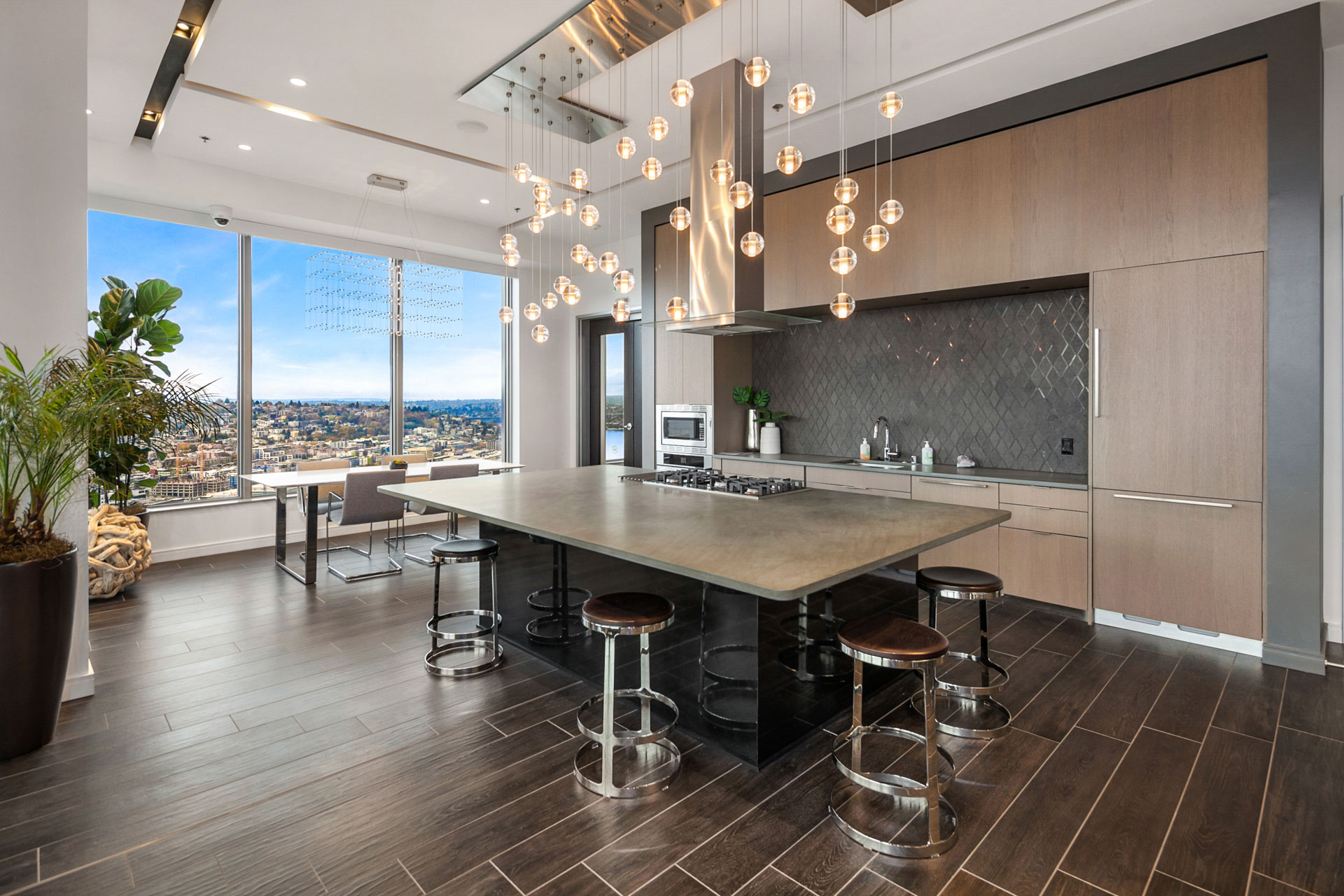 Modern kitchen featuring a large island with barstools, pendant lights, stainless steel appliances, and flat-front wood cabinetry. The room boasts floor-to-ceiling windows with a view of a cityscape, light wood flooring, and a dining area with a table and chairs.