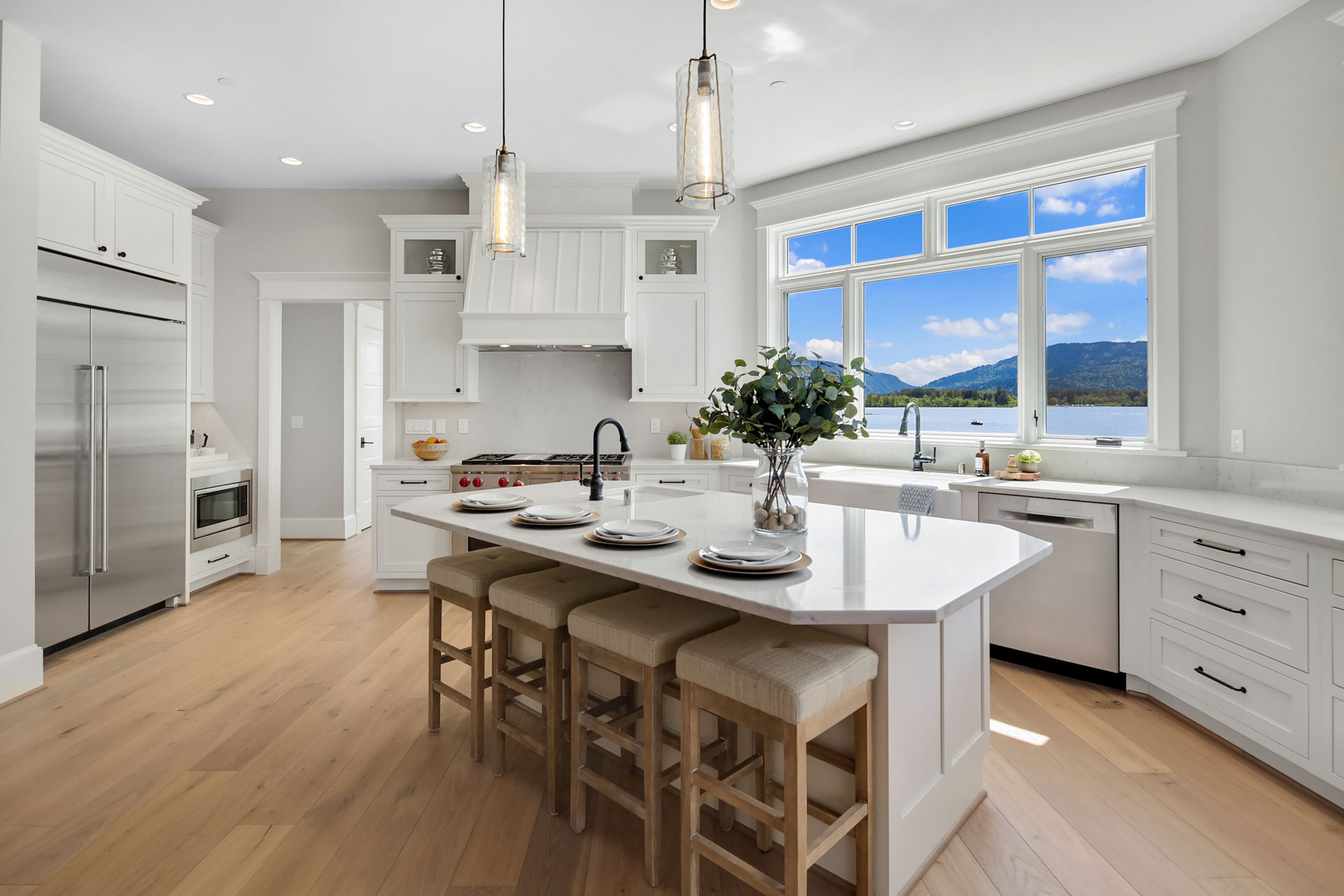 A modern kitchen with white cabinets, stainless steel appliances, and a large island featuring a white countertop and four stools. Three pendant lights hang over the island, while a window offers a scenic view of a lake and mountains. The floor is light wood.