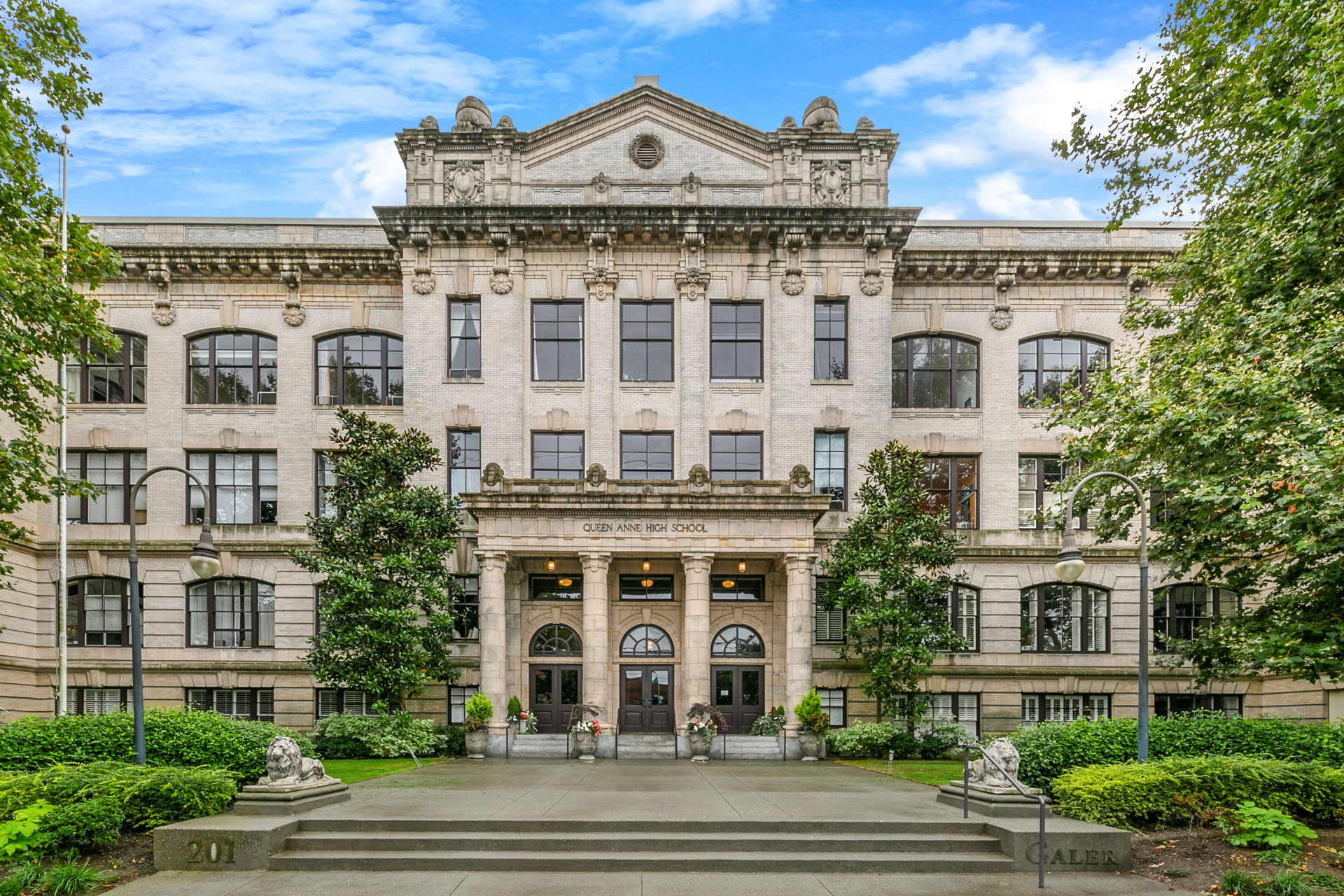 A grand, old-style building with detailed architectural features and columns. The sign "Queen Anne High School" is visible above the main entrance. The entrance has three arched doorways and a few steps leading up. Lush greenery flanks each side of the path.