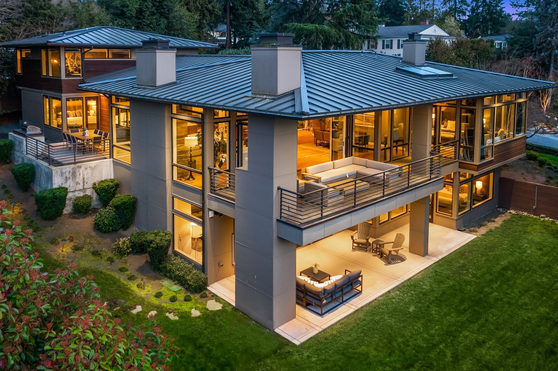 A modern, two-story house with large windows and a flat roof, illuminated warmly from the inside. The exterior features a spacious outdoor patio with seating areas on the lower and upper levels. The surrounding landscape includes well-maintained lawns and shrubs.