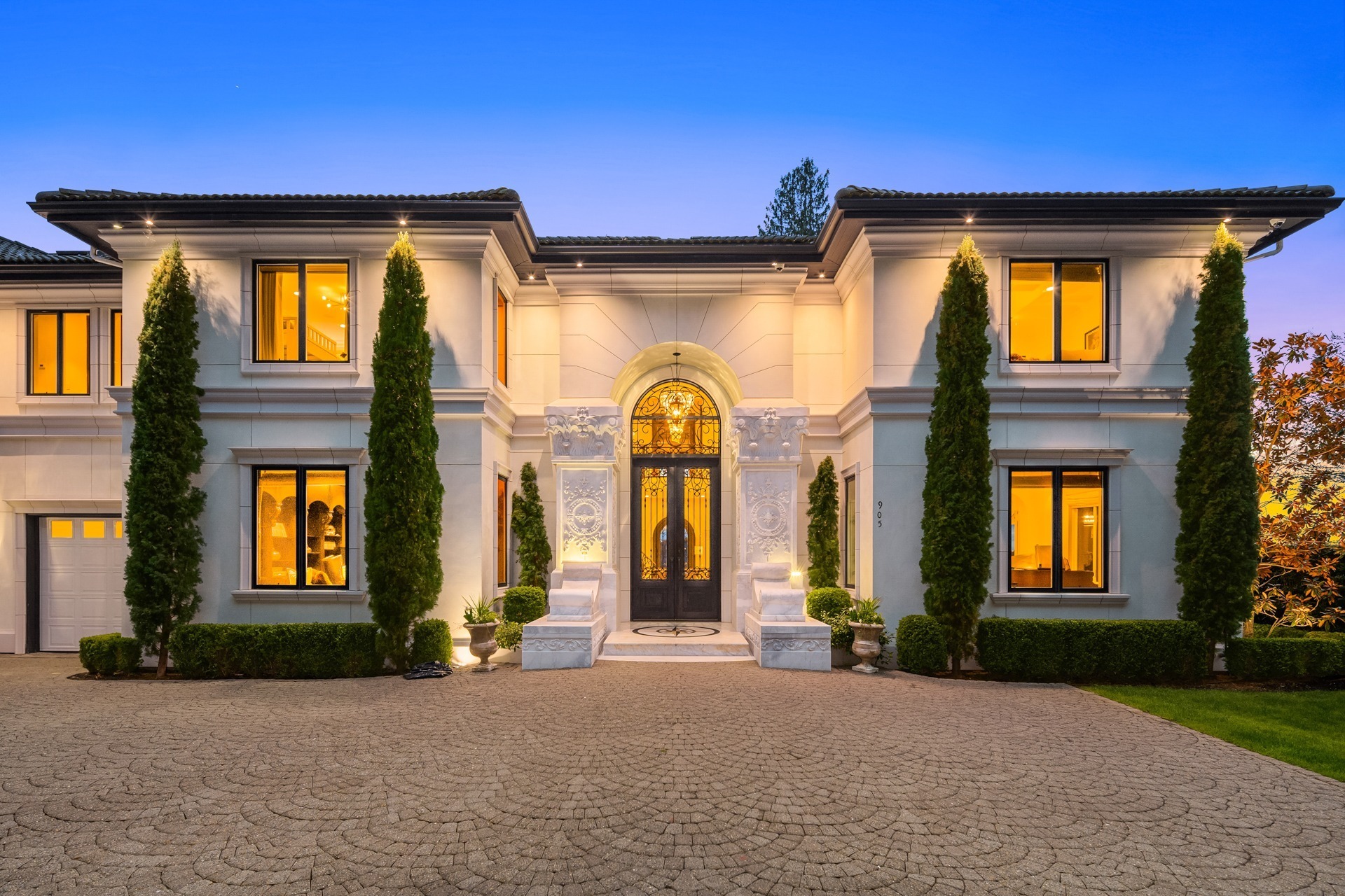 A grand, two-story luxury home with a symmetrical white facade, large arched entrance, intricate iron double doors, and tall windows, illuminated by warm exterior lighting. The driveway is paved with brick, and there are tall, narrow trees flanking the entrance.