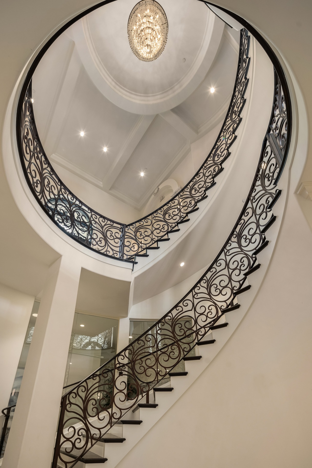 A grand, spiral staircase with ornate, black wrought-iron railings and white walls ascends under a round ceiling. A chandelier hangs overhead, illuminating the elegant architectural design.