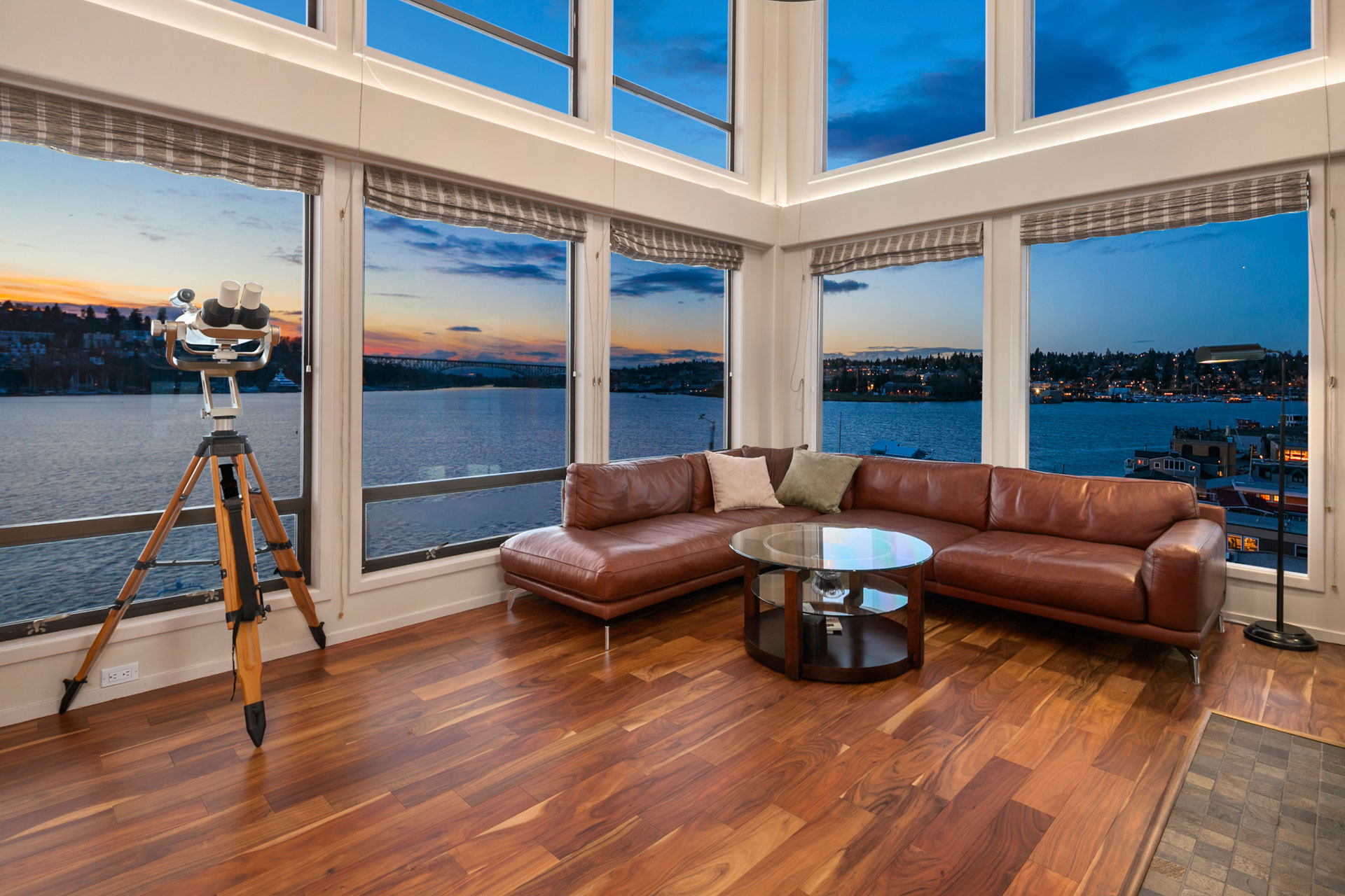 A modern living room with large windows offers a panoramic view of a serene lake at sunset. The room features a brown leather sectional sofa with a round glass coffee table in front of it. A high-end telescope is positioned by the window, and the floor has rich wooden planks.