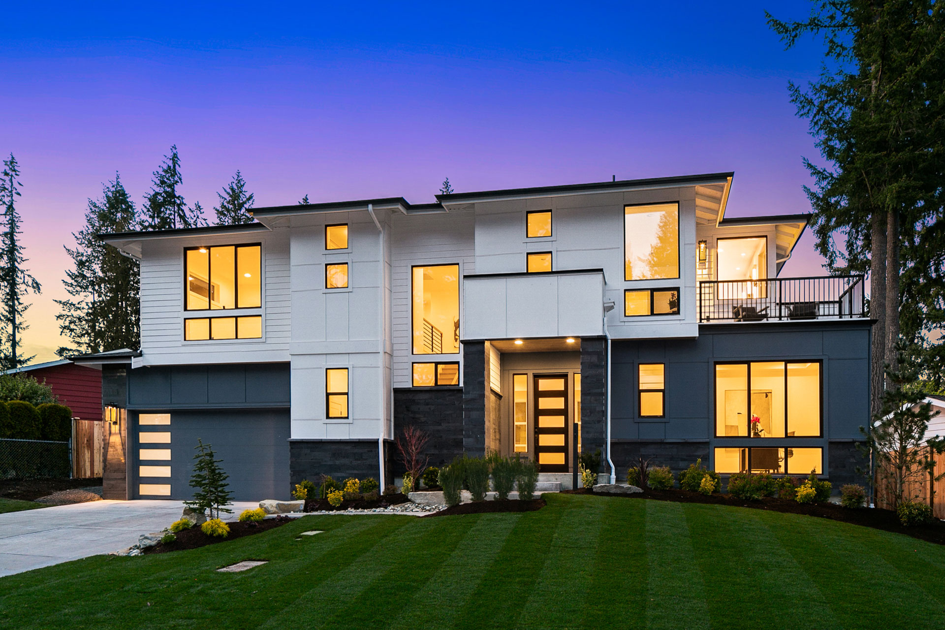 A modern two-story house with a well-manicured lawn and a paved driveway. The house features large windows, a contemporary design with a mix of white and dark exterior materials, and a prominent front entrance. Tall trees and a twilight sky are in the background.