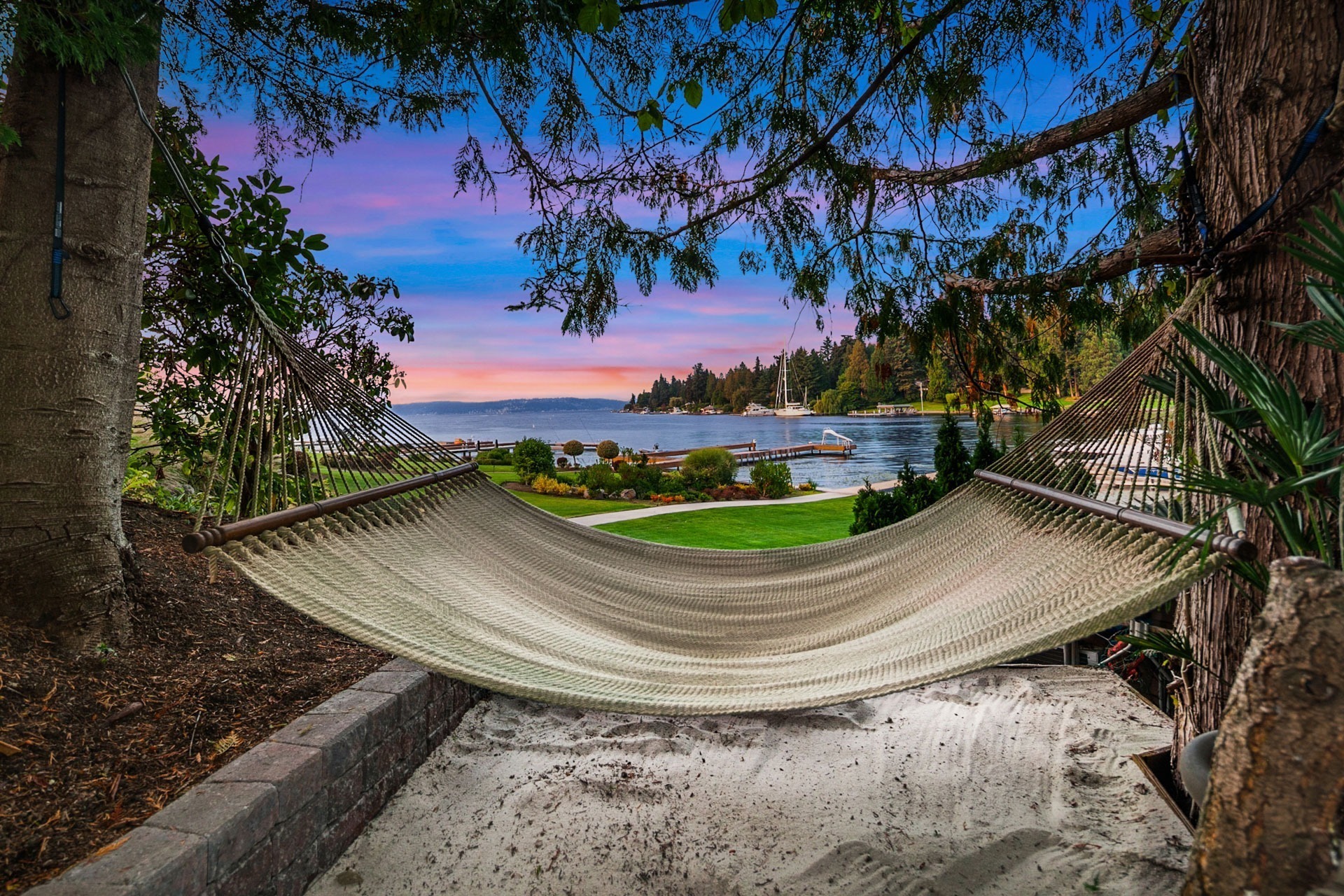 A woven hammock is tied between two trees, overlooking a serene lake during sunset. The sky displays shades of pink and purple. The foreground features a sandy area and the background shows a well-maintained lawn, trees, and a dock extending into the water.