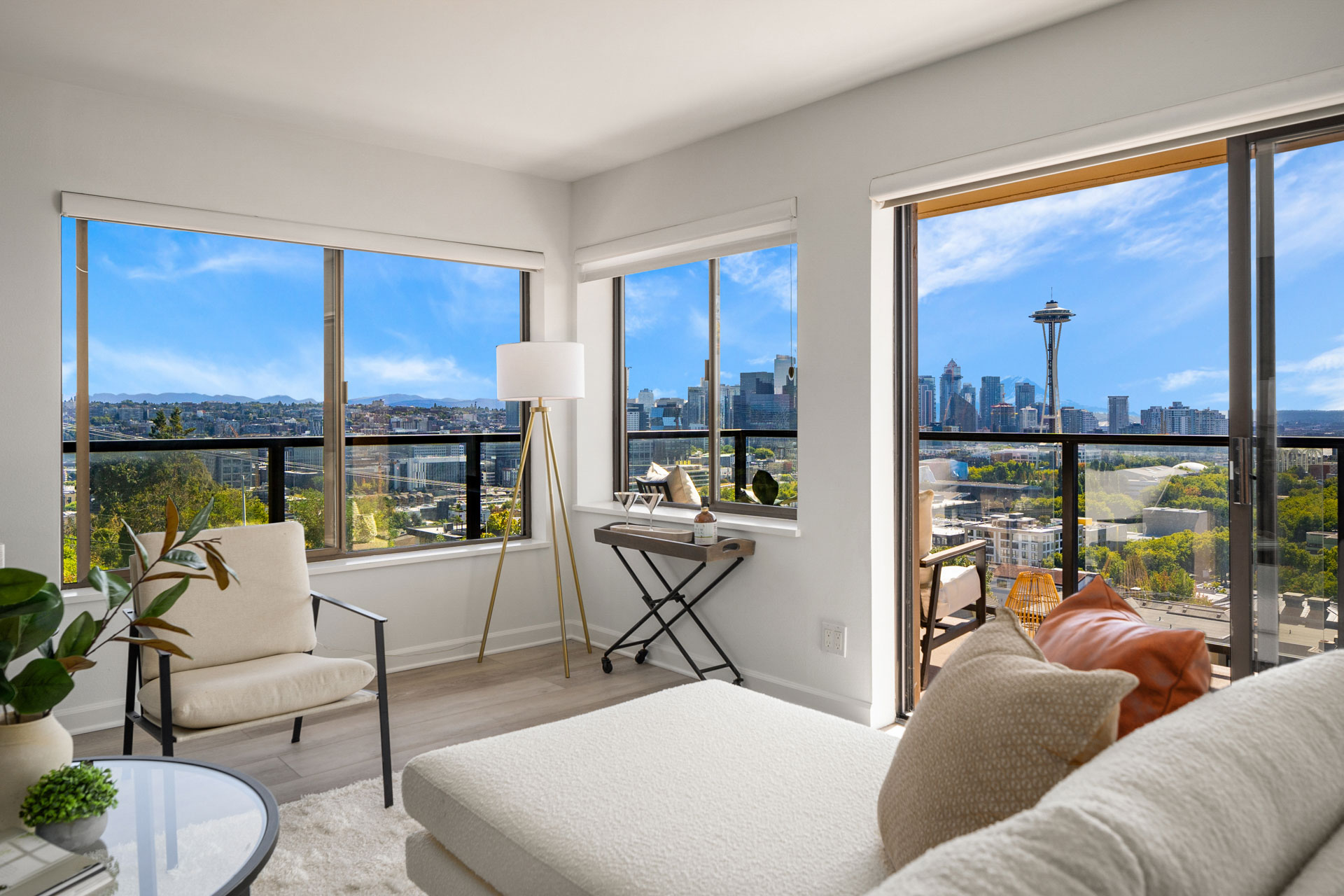 A modern living room with large windows offering a panoramic view of a cityscape, including the Space Needle. The room features a white sofa, a stylish floor lamp, a round coffee table with plants, and a small desk beside one of the windows.