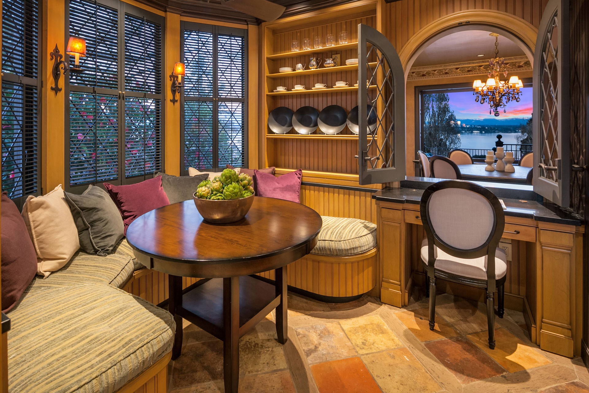 A cozy corner with a round wooden table surrounded by cushioned bench seating and pillows. Adjacent is a small desk with a chair. Shelves with decorative plates line the walls. A large window overlooks a scenic sunset view over water, illuminating the room.