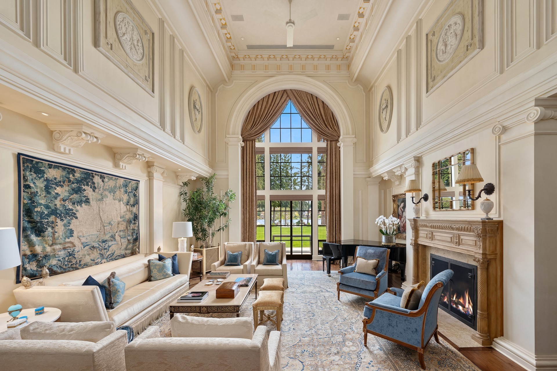 A luxurious living room with high ceilings, ornate wall details, and large windows offering a view of greenery outside. The room features elegant furniture including cream sofas and blue armchairs, a grand fireplace, a piano, and a tapestry hanging on the wall.