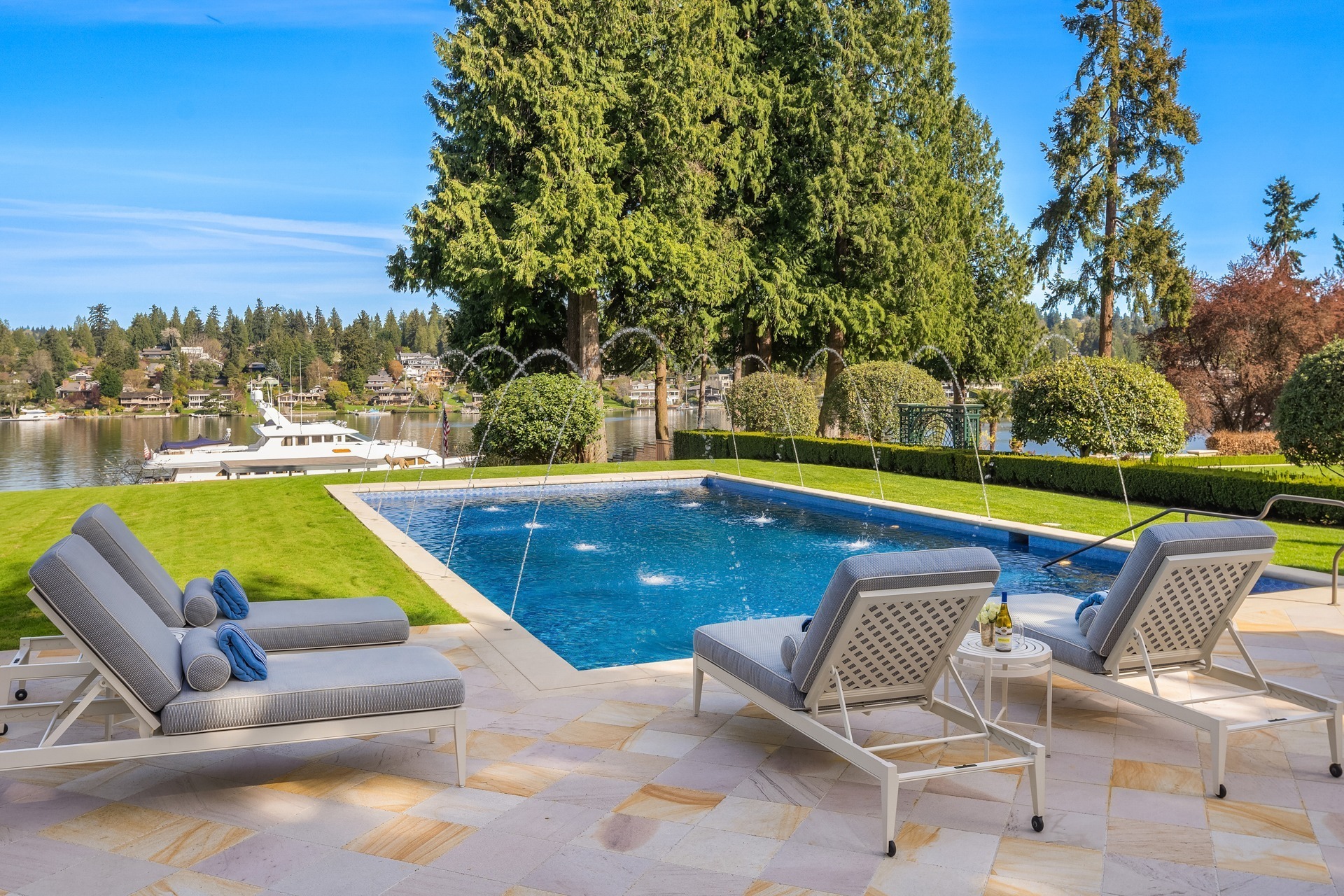 A luxurious outdoor pool area featuring lounge chairs with cushions, set on a stone patio. The pool has water fountains spraying into it and is surrounded by manicured lawns, trees, and a scenic view of a lake with boats in the background.