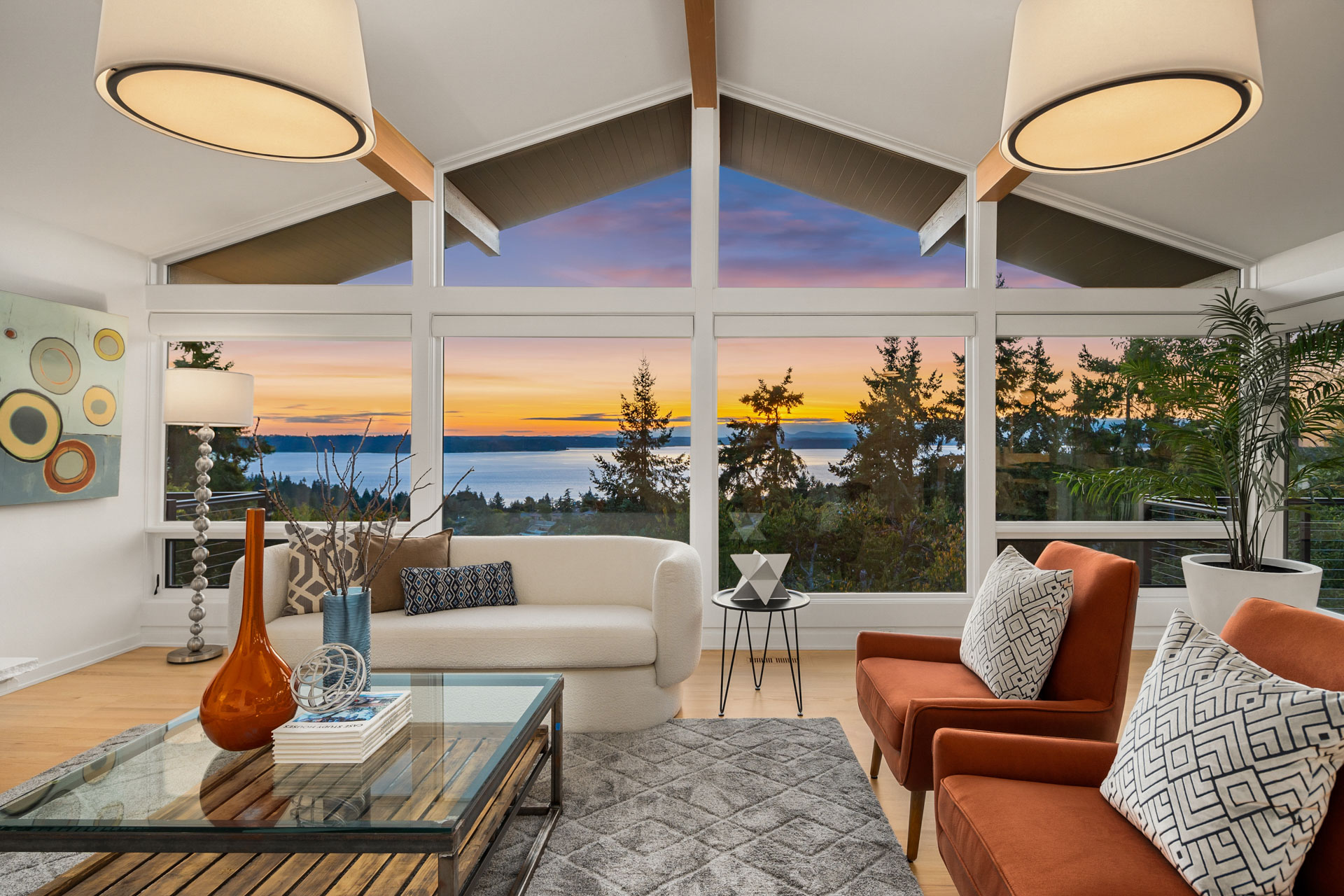 A stylish living room featuring large windows with a stunning sunset view over a body of water and trees. The room includes a white curved sofa, two orange armchairs with patterned cushions, a glass coffee table with decorative items, and modern pendant lights.