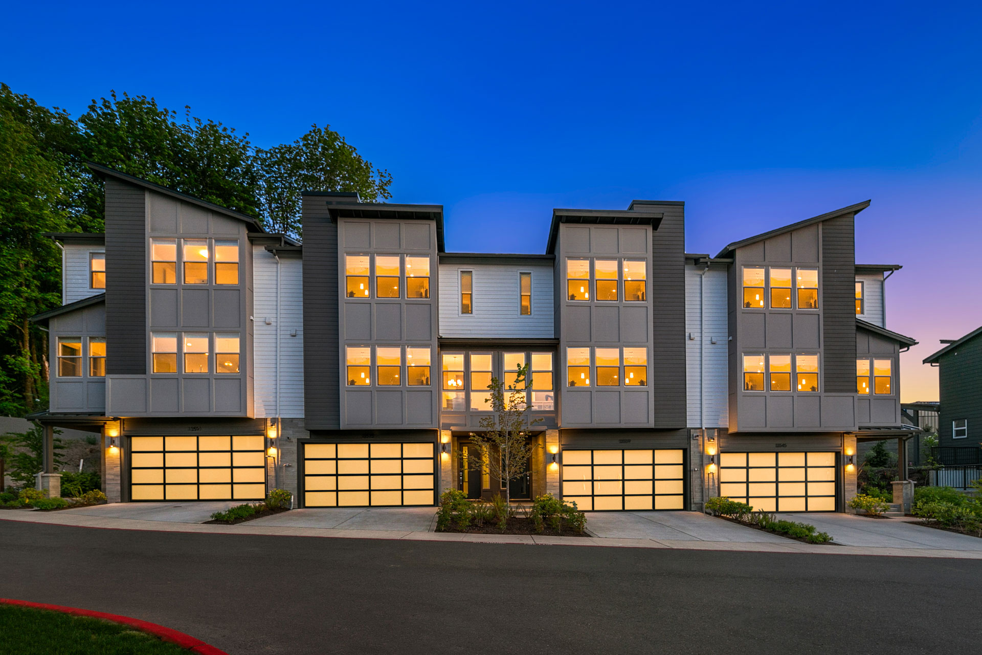 A symmetrical modern townhouse complex with three units, each featuring large windows and individual garages with lit door panels. The buildings have sloped roofs and are illuminated at dusk, emphasizing the contemporary architecture. Lush greenery surrounds the property.