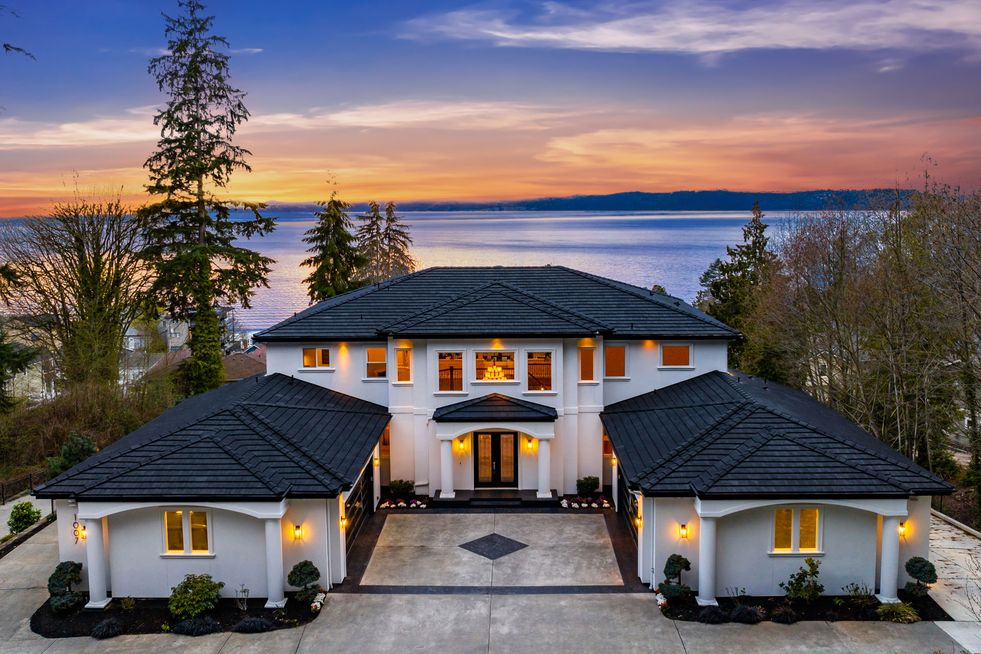 A luxurious white mansion with dark roofing sits against a backdrop of a serene lake and a vibrant sunset sky. The house features a grand entrance with tall columns, multiple windows, and neatly landscaped surroundings. Tall trees frame the sides of the property.