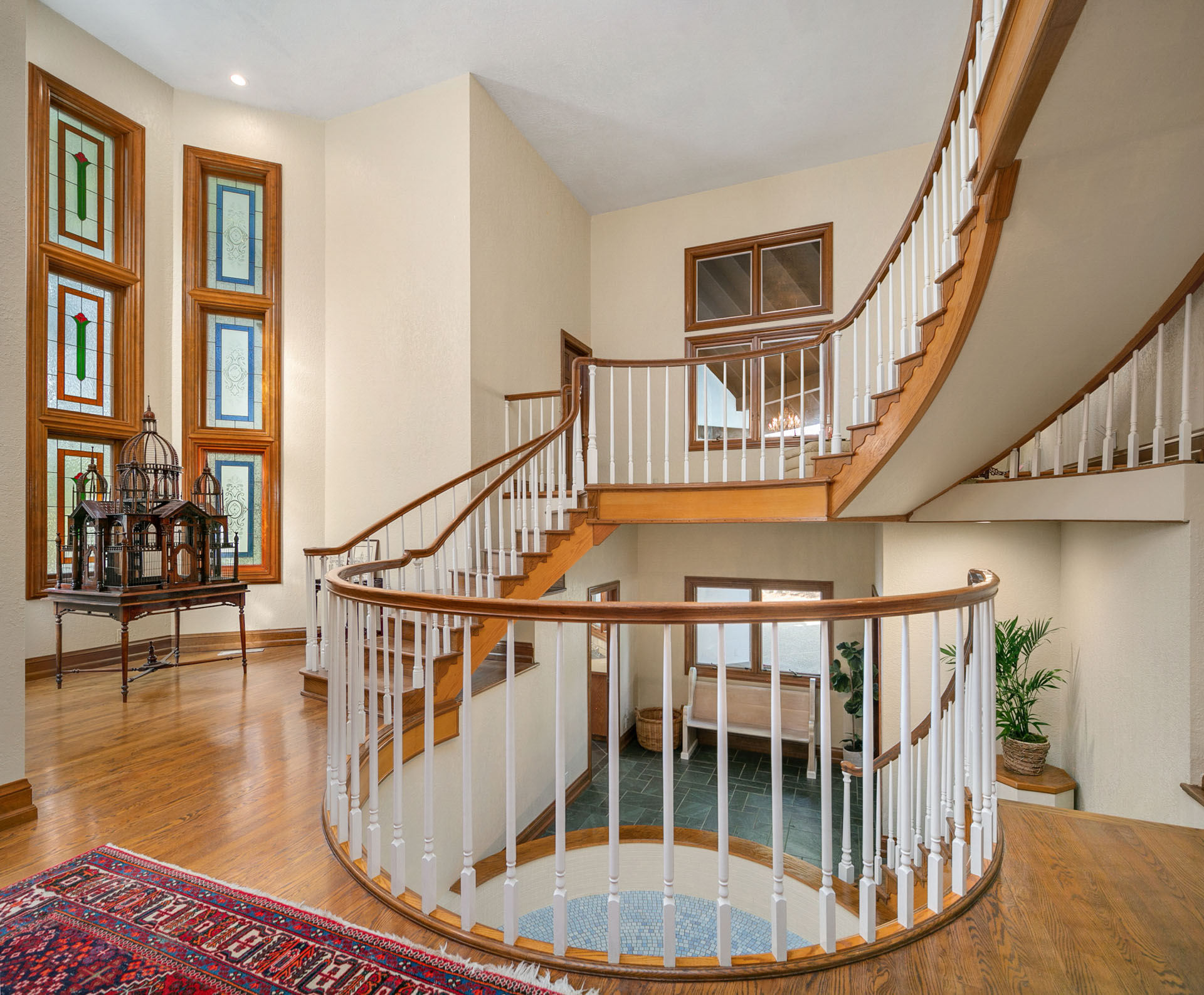 A spacious, well-lit foyer with a grand, curved staircase featuring wooden handrails and white balusters wraps around an open area. Decorative stained-glass windows adorn one wall, and a red-patterned rug covers part of the hardwood floor.