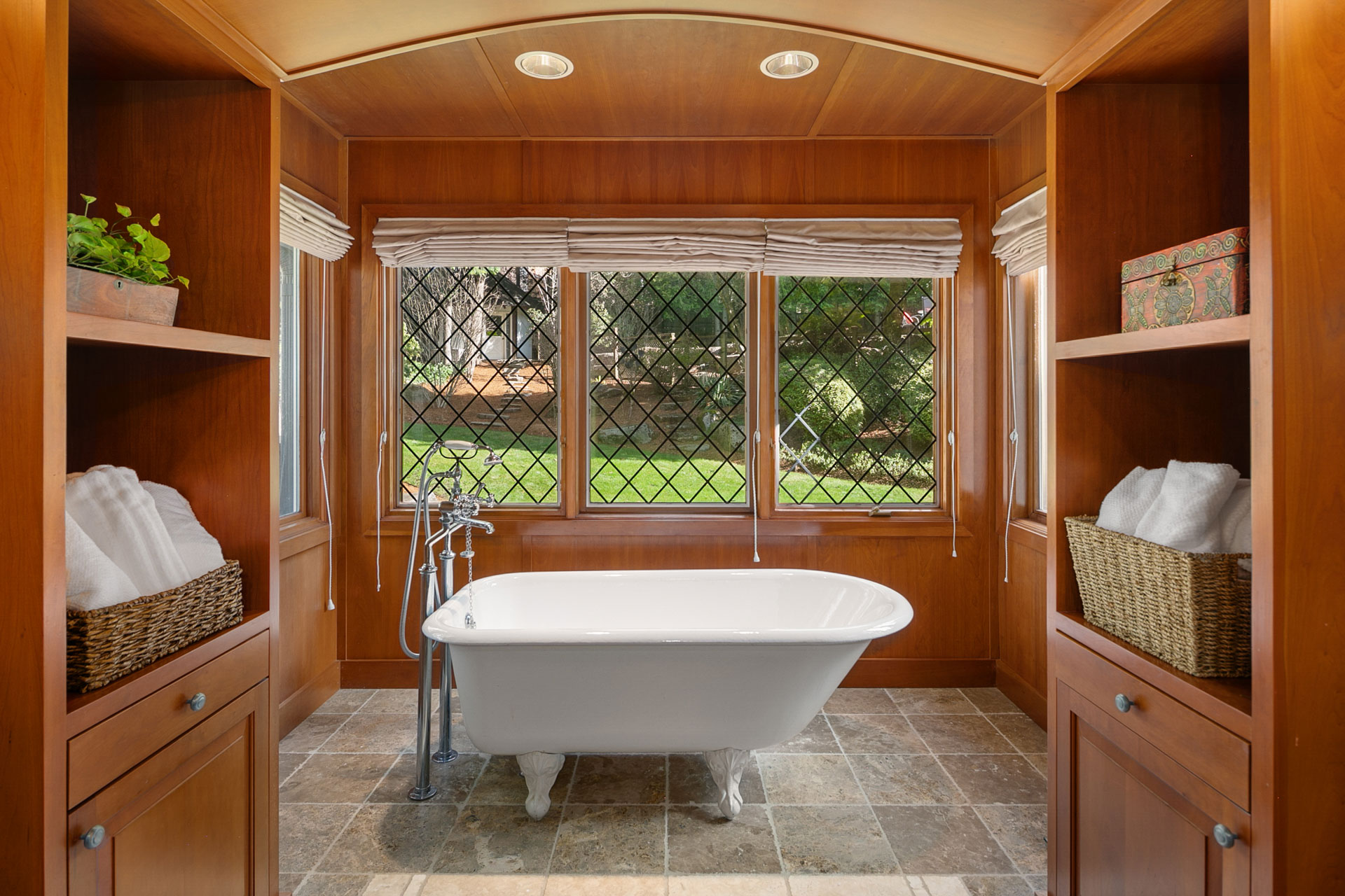 A luxurious bathroom with wooden walls and shelves, featuring a white clawfoot bathtub placed in front of large diamond-paned windows. The shelves are adorned with wicker baskets and a potted plant. The floor is tiled, complementing the warm and inviting ambiance.