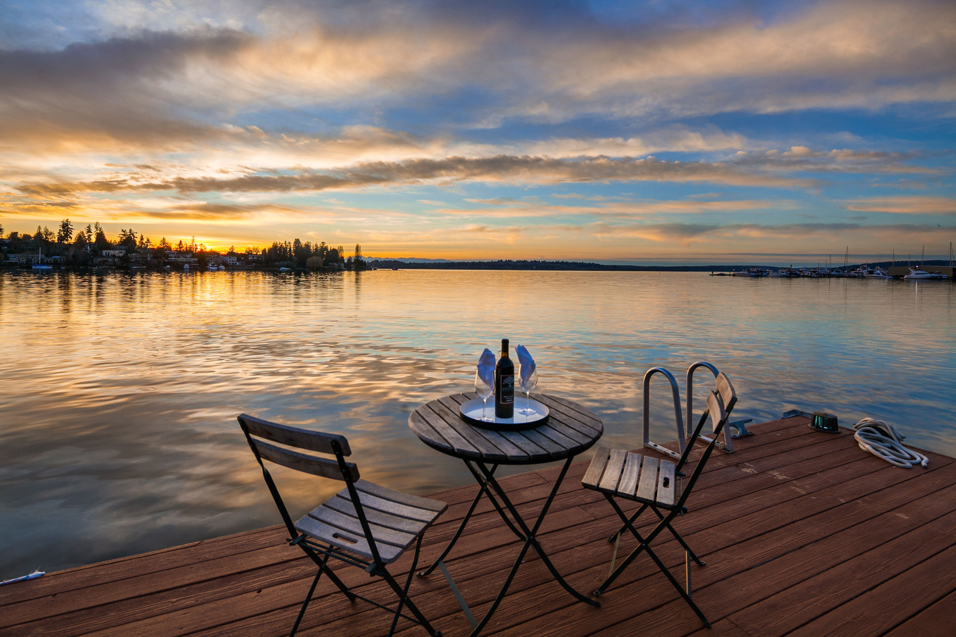 A serene lakeside scene at sunset with a wooden deck featuring a small round table set with a bottle of wine, glasses, and a wine cooler between two folding chairs. Calm waters reflect the golden and blue hues of the sky, with trees and houses visible in the distance.