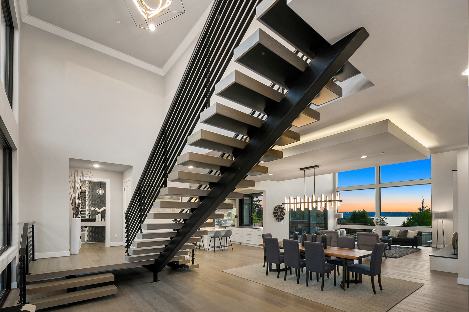 A modern open-concept living space featuring a large floating staircase with wooden steps and black metal railings. The dining area has a table set for eight. The living area is furnished with sofas and a large window showcases a sunset view.