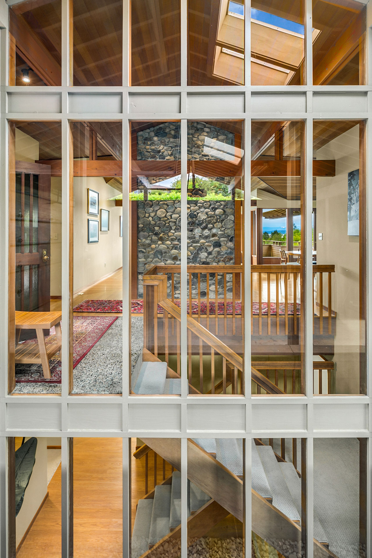 View through large window panes into a modern home with wooden interiors. The image reveals a staircase leading up to a room with stone accent walls and skylights, framed art on the walls, and a glimpse of a scenic landscape in the background.