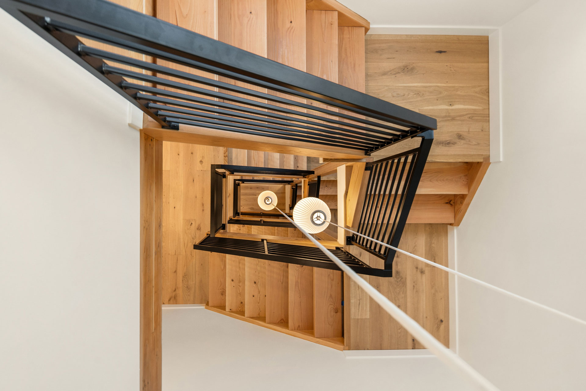 Overhead view of a modern, wooden spiral staircase with black railings. The stairs twist around a central axis, and a white pendant light hangs down the center. The photo captures the geometric patterns and warm tones of the wood, creating a sense of depth.