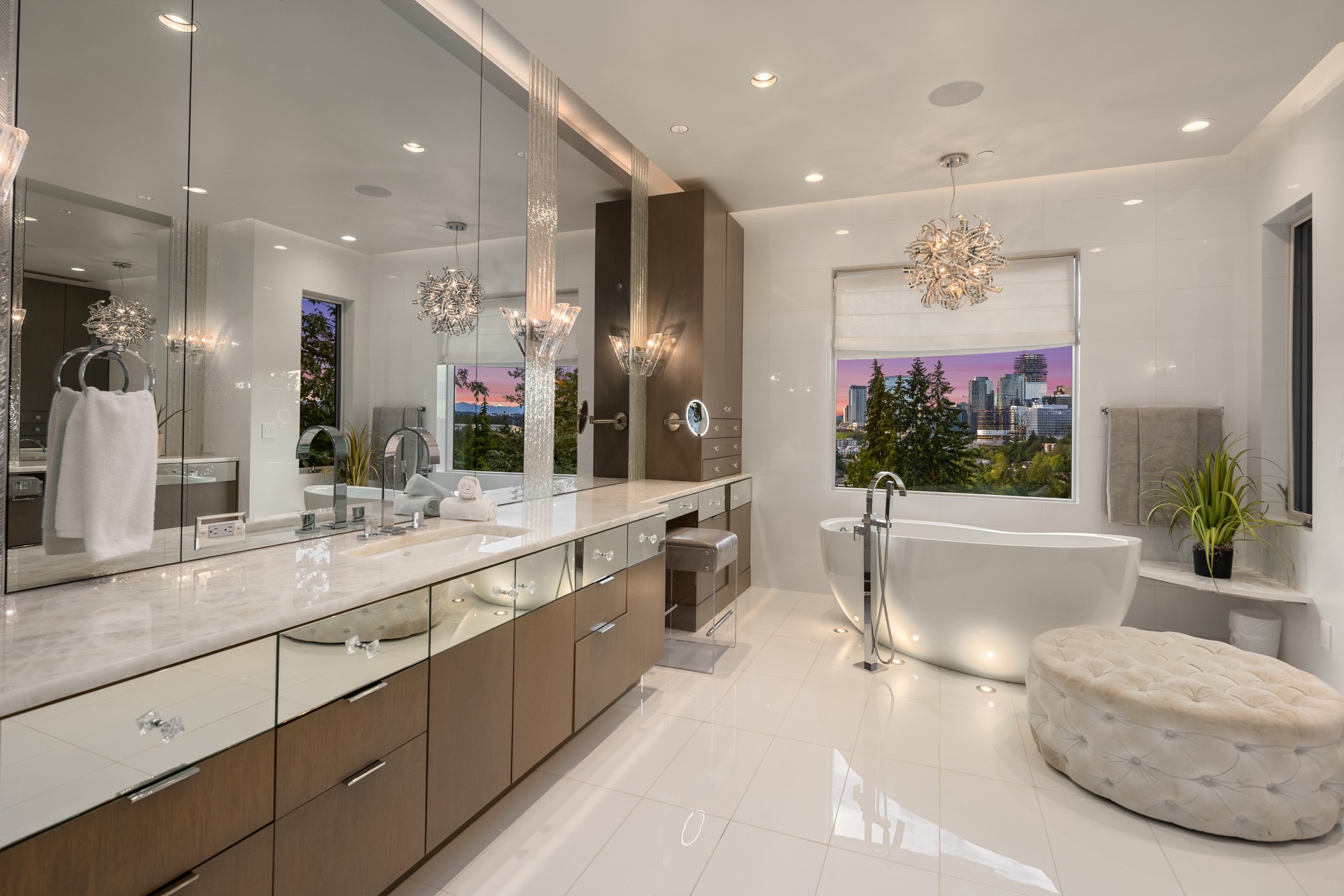 A modern bathroom with a large mirror, double sink vanity, and marbled countertop. A freestanding bathtub is positioned near a window with a cityscape view. The room is adorned with contemporary lighting fixtures and a tufted round ottoman on the polished white floor.