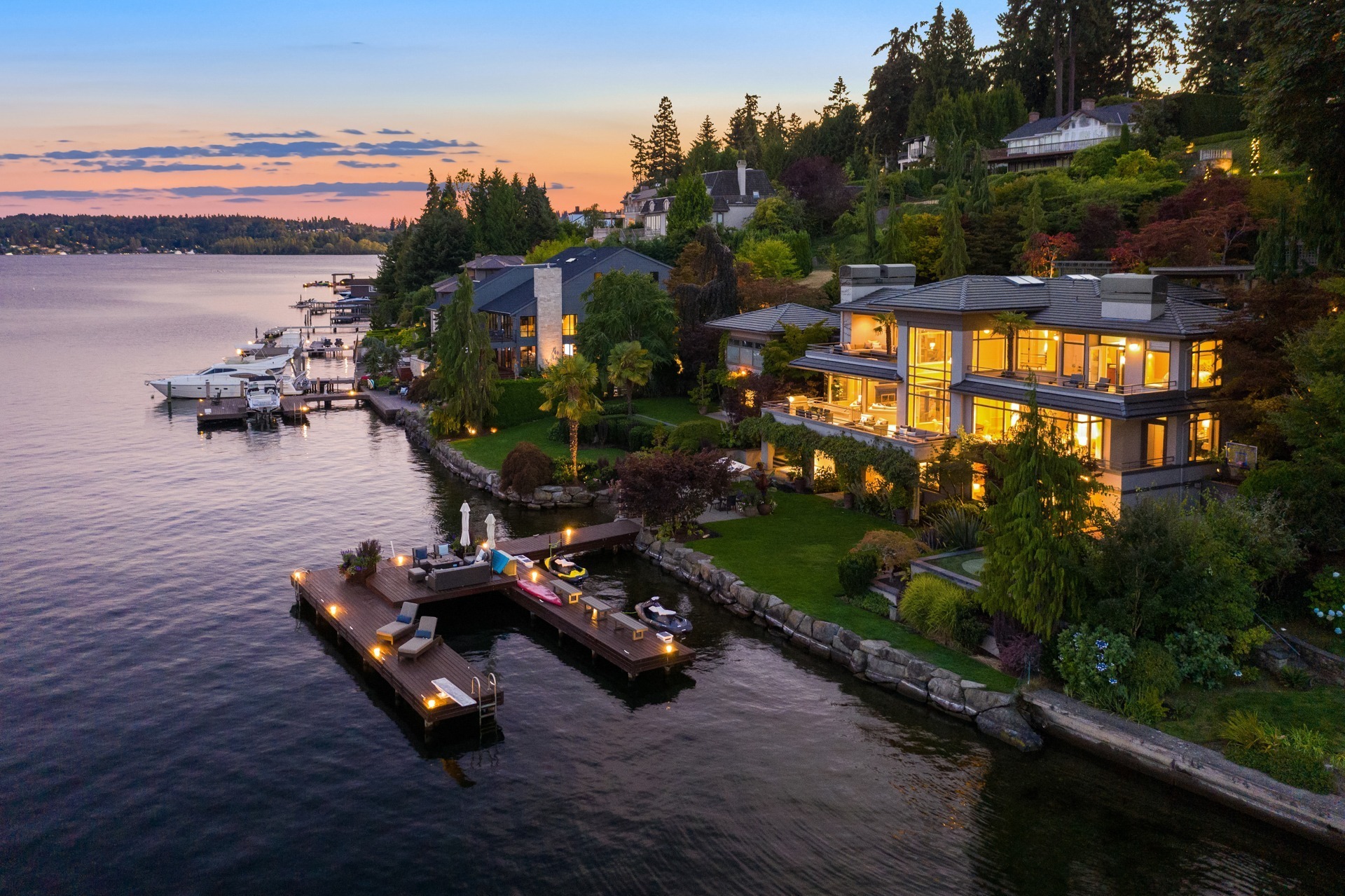 A serene lakeside neighborhood at sunset featuring spacious, modern homes with large glass windows glowing from interior lights. The homes are surrounded by lush greenery and have private docks extending over the water, equipped with boats and seating areas.