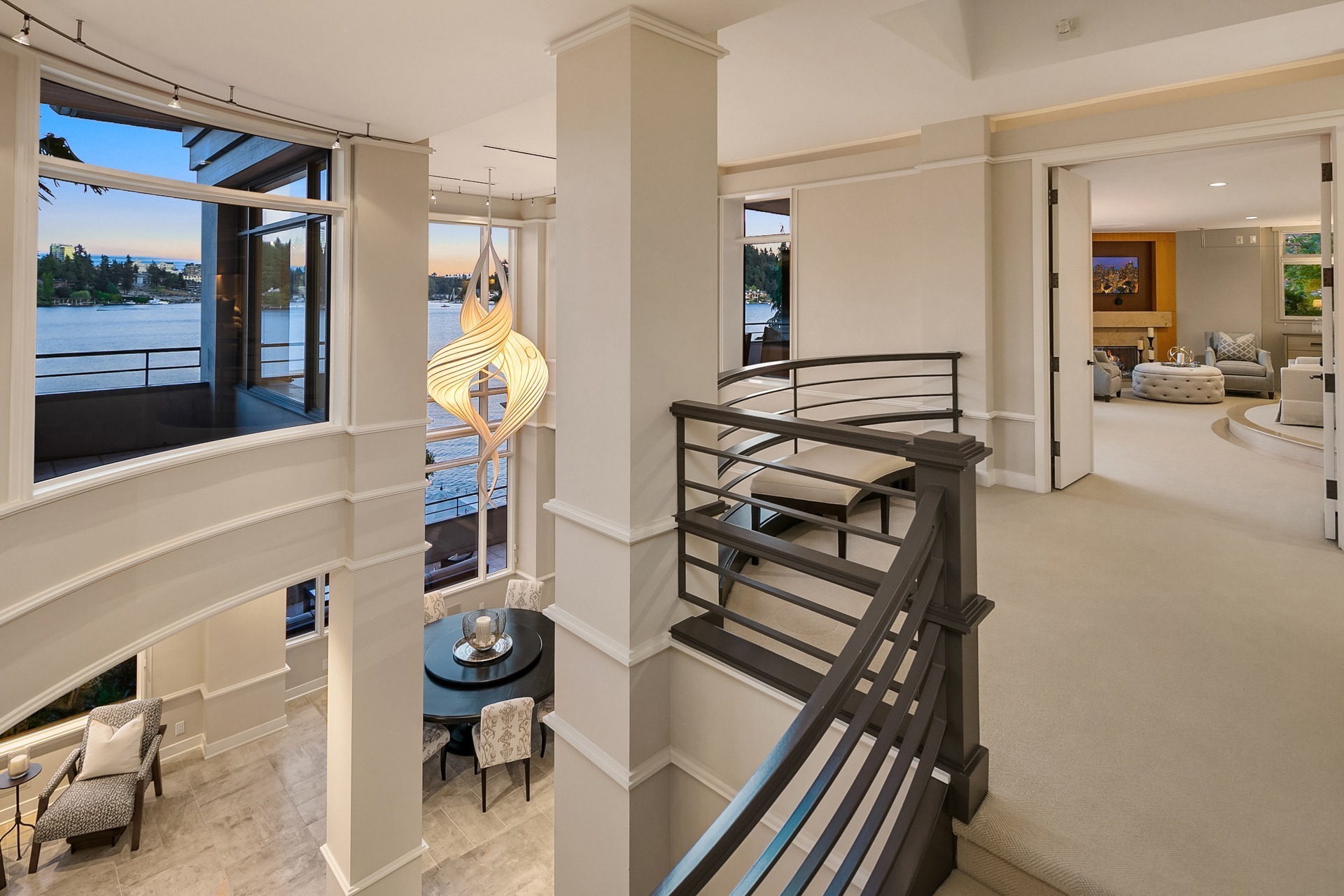 A modern, spacious home interior with a view of a lake through large windows. A staircase with dark railings leads from the lower dining area to an upper level. A unique pendant light hangs above the dining table set with chairs. The upper level features a cozy living area.