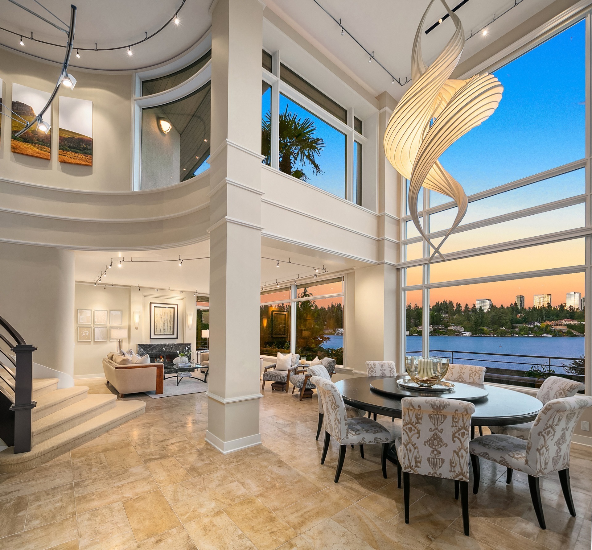 Spacious modern living room with large windows offering a waterfront view at sunset. Features beige tiled floors, round dining table with patterned chairs, curvy hanging sculpture, two-story ceiling, staircase, and a cozy sitting area with contemporary decor.