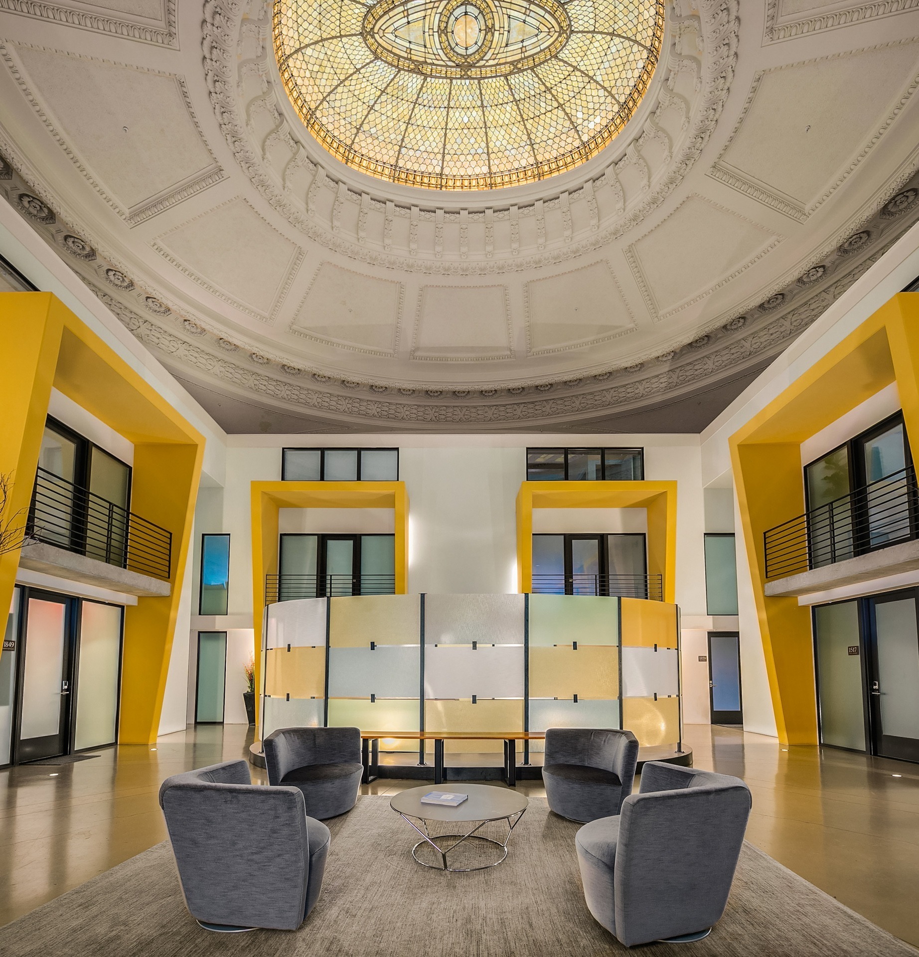 A modern lounge area is shown featuring four gray chairs around a small table on a light-colored rug. The space is adorned with vibrant yellow walls, large two-story windows, and an ornate domed ceiling with a detailed, illuminated design.