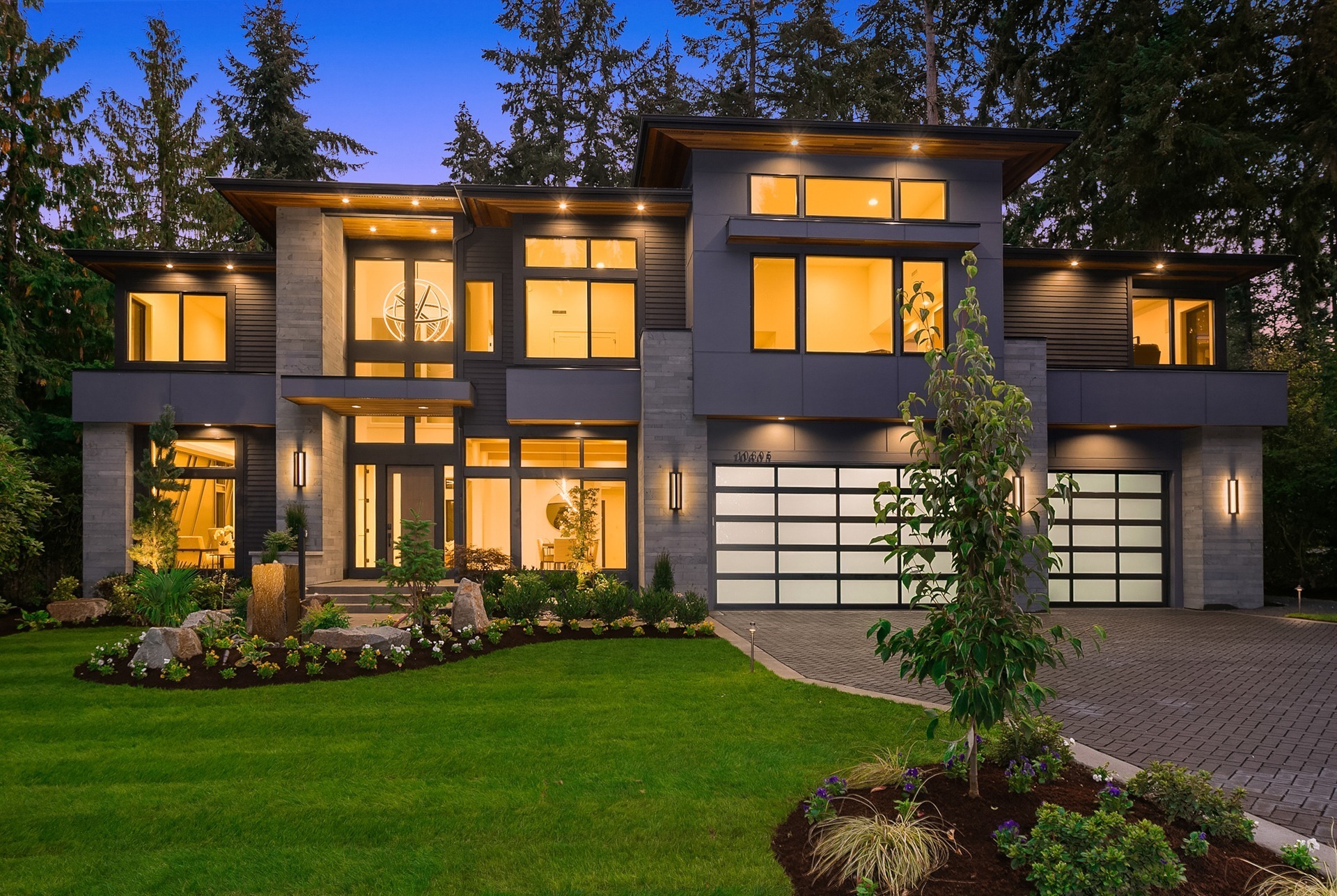 Modern two-story house with large windows, exterior lights, and a well-manicured lawn. The entrance is lit warmly, and a tree stands in the front yard. The spacious driveway leads to a garage with a frosted glass door. The backdrop is a serene forest.