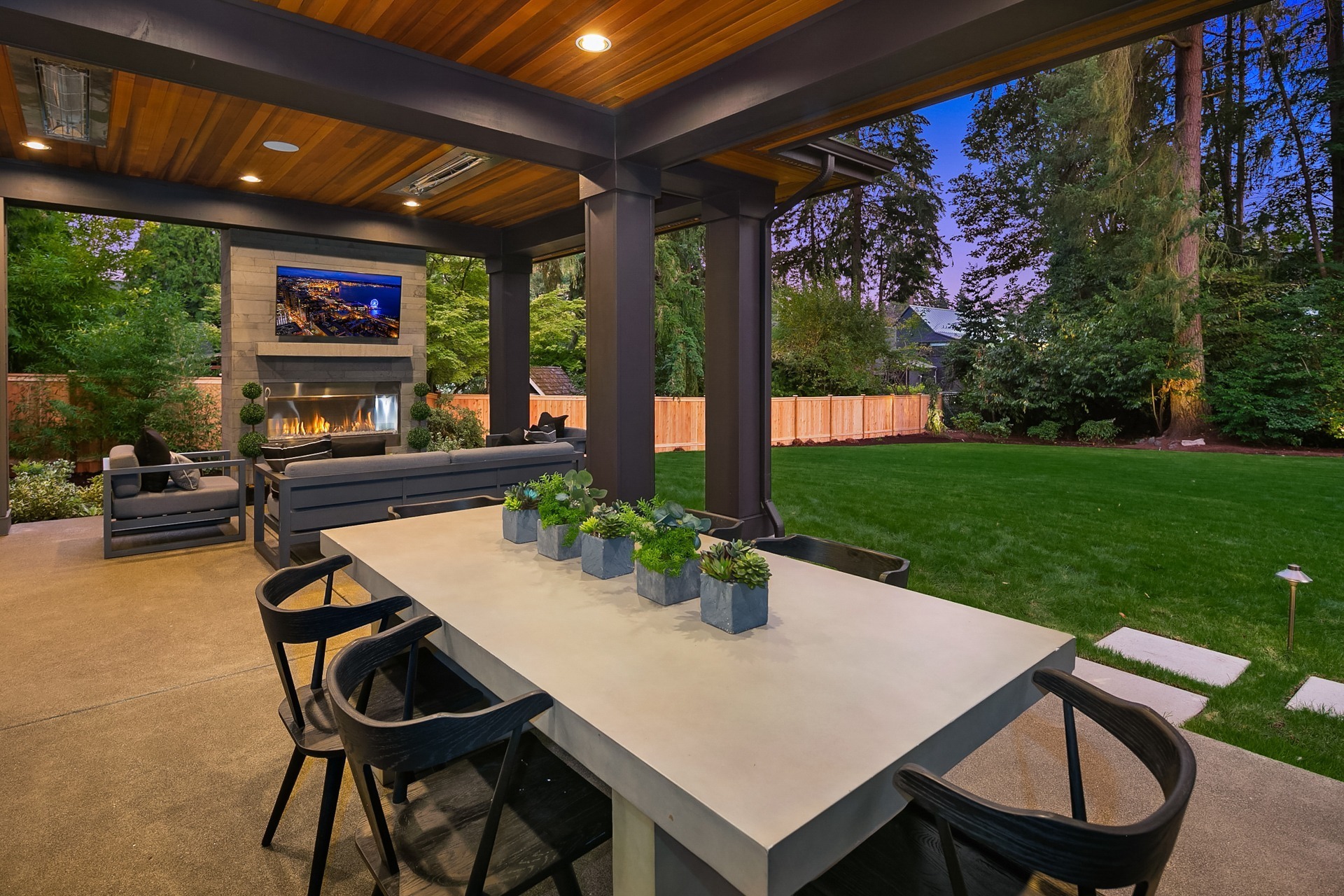 A modern outdoor patio features a dining table with black chairs, potted plants, and an adjacent lounge area with a fireplace and TV. The patio has a wooden ceiling with recessed lighting and opens to a spacious, well-manicured lawn surrounded by trees and a fence.