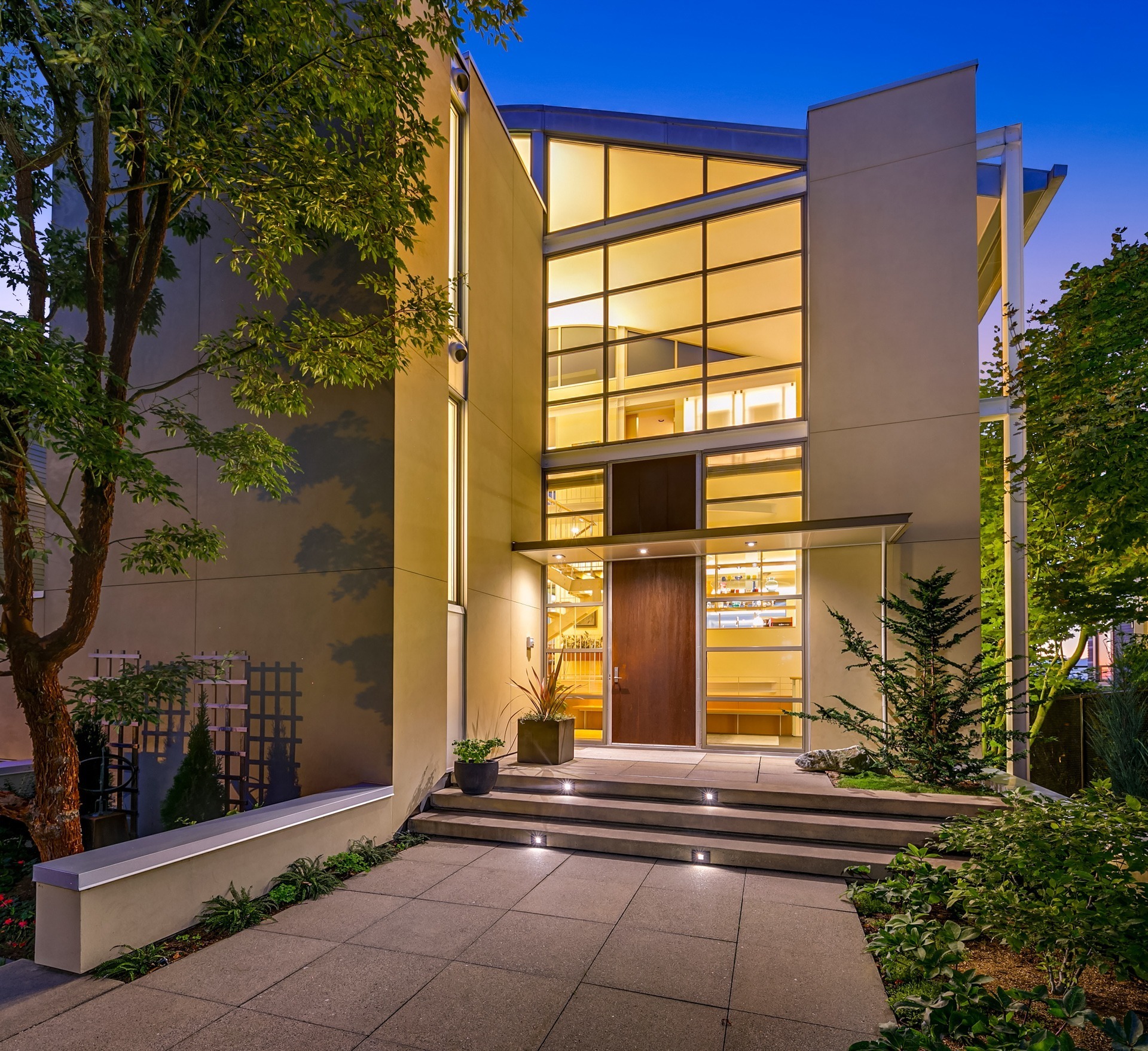 A modern, two-story house entrance is shown at dusk. The house features large, multi-pane windows and a minimalist design with clean lines. Steps lead up to a wooden front door. Trees and plants surround the walkway, enhancing the contemporary aesthetic.