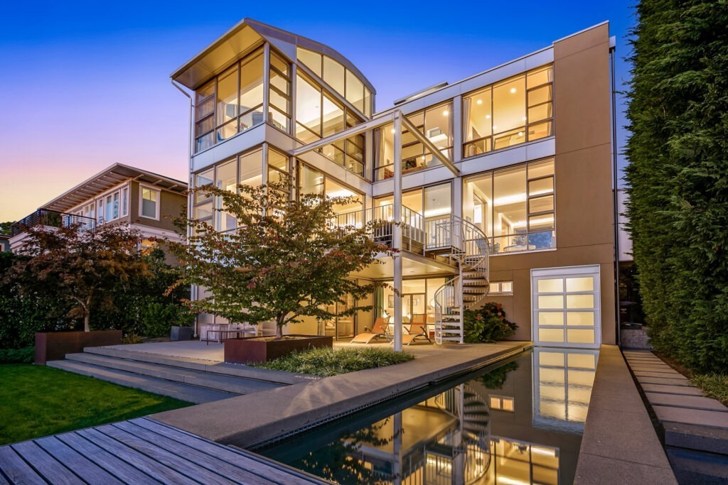 A modern three-story house with large glass windows and a sleek, geometric design is illuminated at dusk. A narrow reflecting pool runs parallel to the house, and a spiral staircase connects the upper levels. Lush greenery surrounds the property.