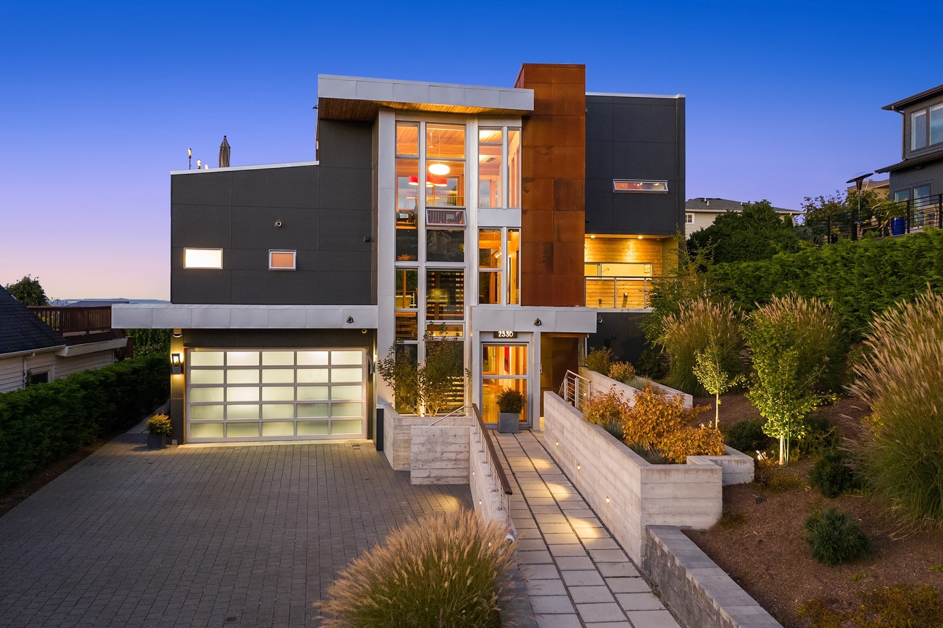 A modern, multi-story house with large glass windows, metal and wooden accents, and a flat roof. The entrance features a well-lit pathway with greenery on both sides. The sleek design is complemented by outdoor lighting and a paved driveway.