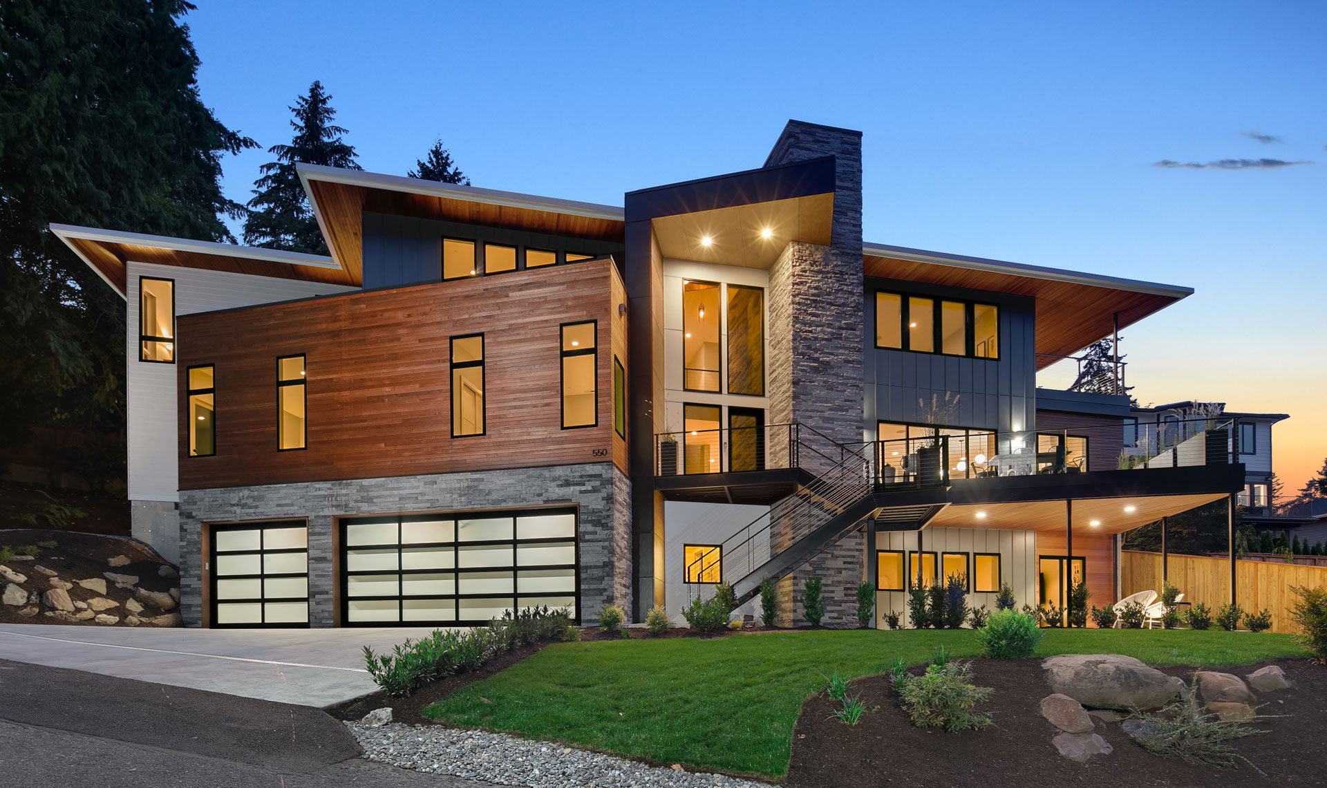 Modern two-story house with wood and stone exterior features large windows, a spacious balcony, and a three-car garage with glass doors. The house is illuminated, highlighting its architectural design, with neatly landscaped surroundings and a twilight sky.