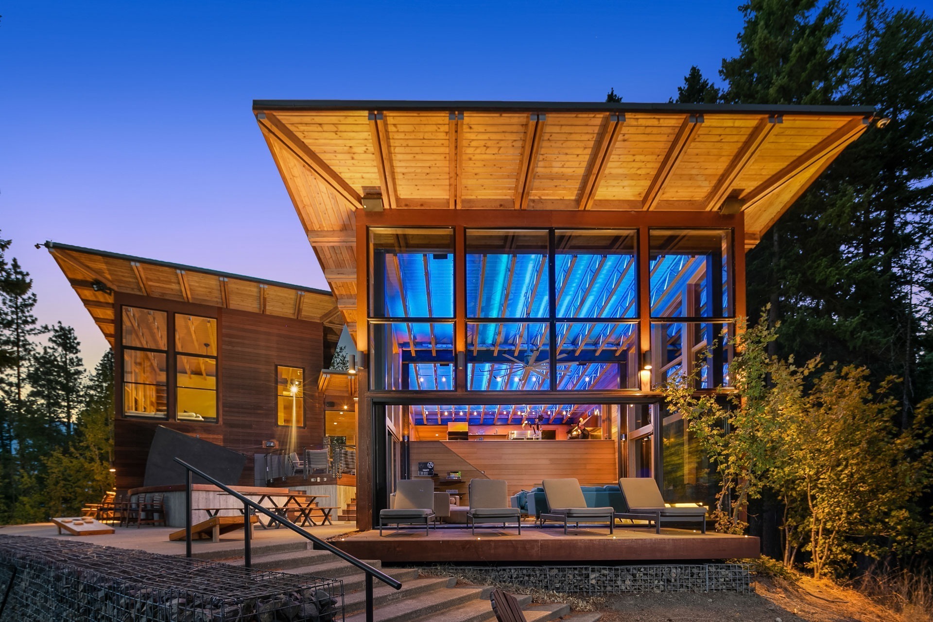 Modern house with large glass windows and wooden accents, featuring an overhanging roof structure illuminated by blue lighting. The outdoor area includes a seating space with lounge chairs and trees framing the contemporary design. The sky is clear at twilight.