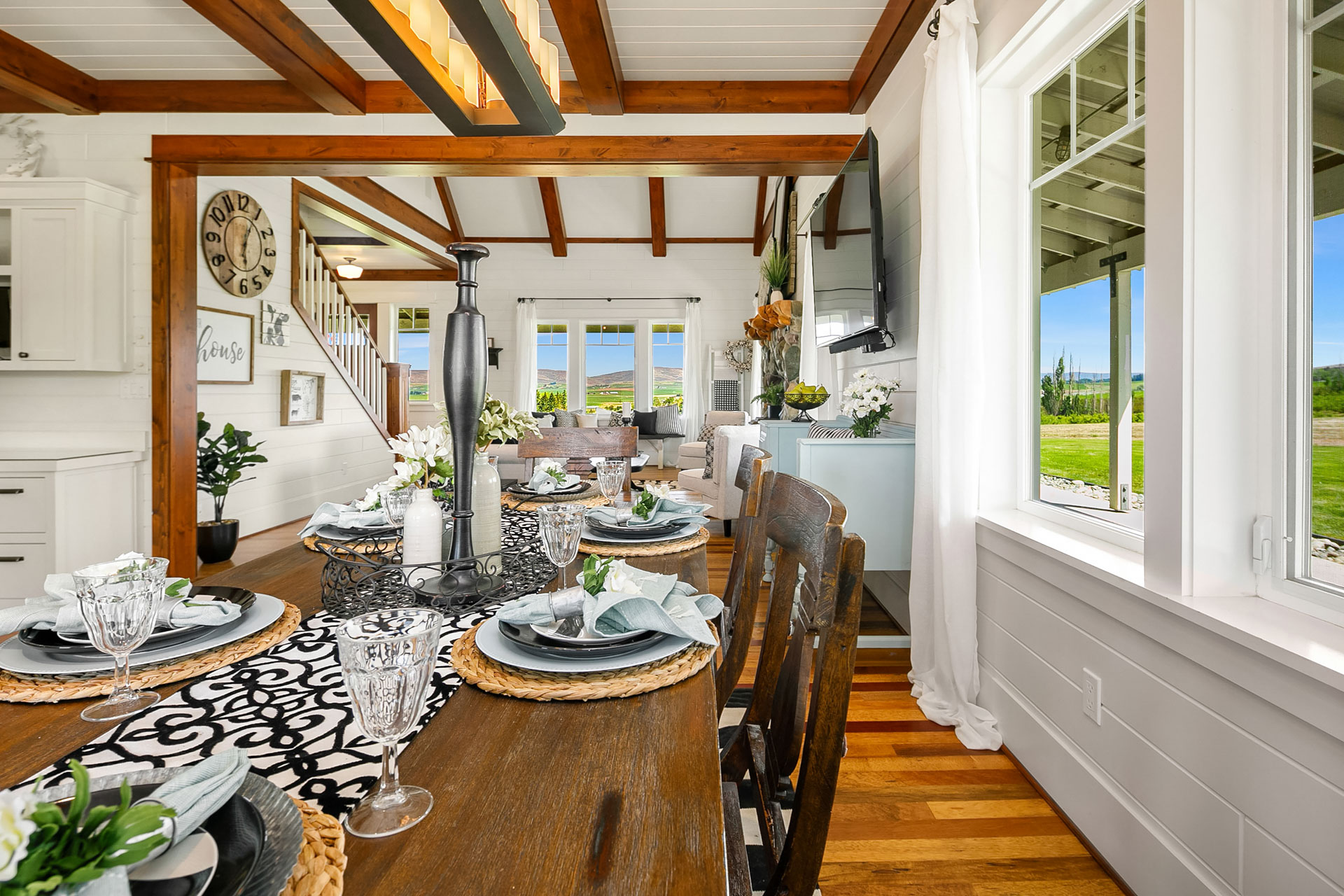 A beautifully set dining table in a bright room with white walls and wooden beams. The table is adorned with gray and white dishes, glassware, and floral arrangements. Large windows offer a scenic view of greenery outside, and elegant decor completes the interior.