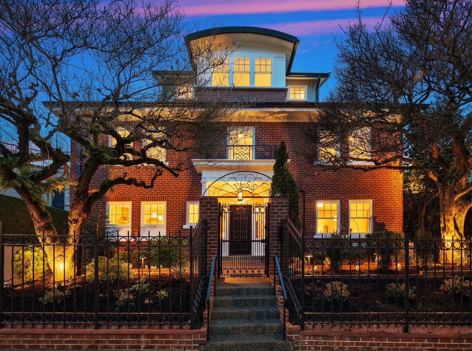 Charming two-story brick house at dusk with warm lights illuminating the windows. The house features a gated entrance, manicured garden, and trees framing the path to the front door. The sky displays vibrant shades of purple and pink.