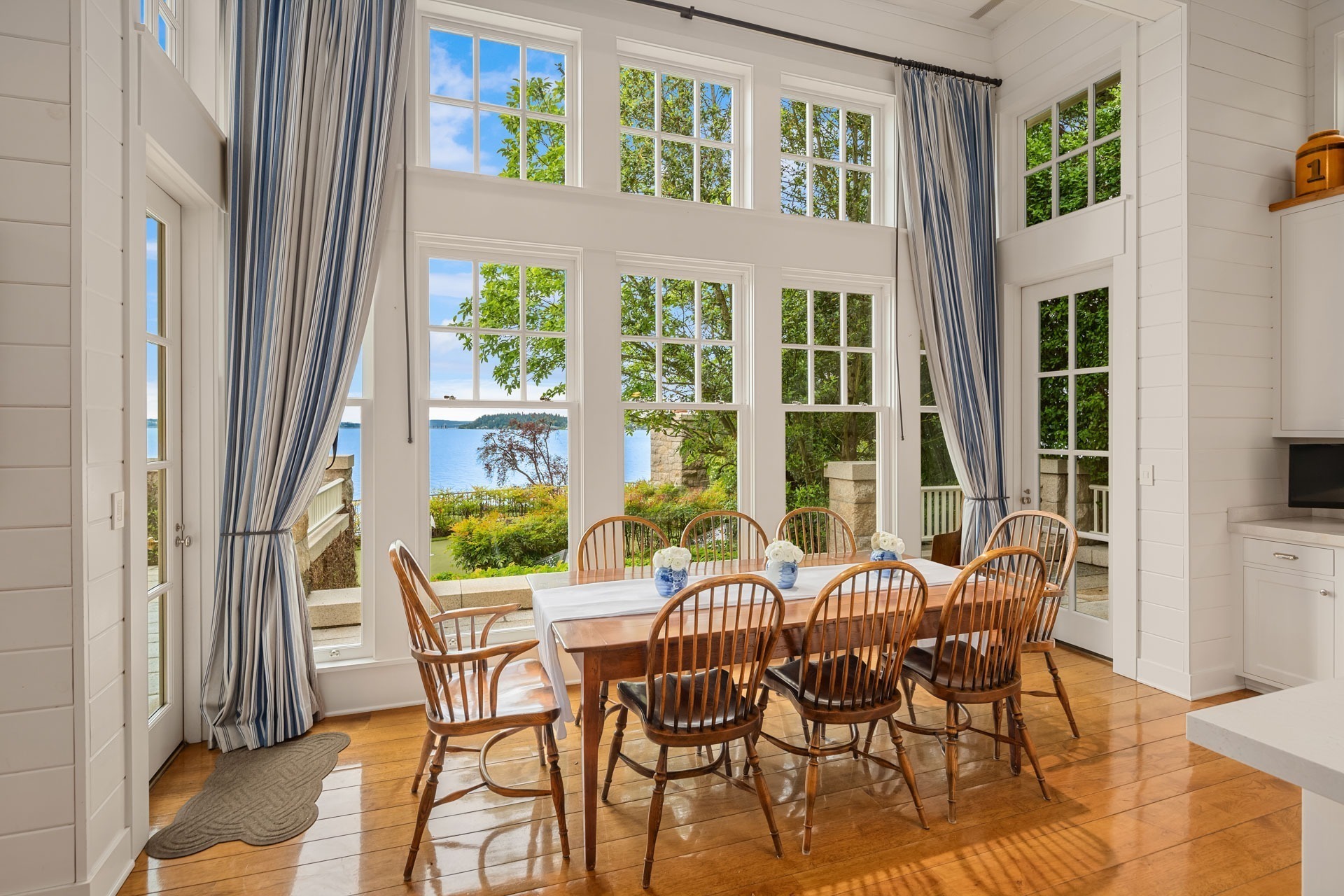 A dining area with large windows offering a scenic view of a body of water and trees outside. The space features a wooden table surrounded by eight wooden chairs, a blue and white tablecloth, and striped blue curtains framing the windows.