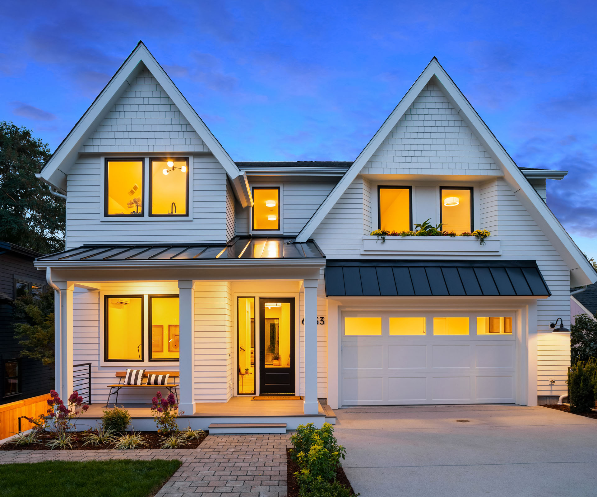 A modern, two-story house with white exterior siding, large windows, and a black metal roof. The front features a porch with chairs, manicured landscaping, and a well-lit driveway leading to a double garage. The sky is twilight blue.