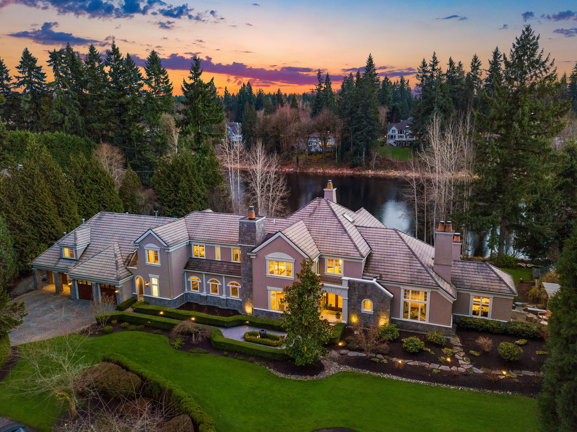 A large, elegant house is nestled amidst tall trees by a serene lake at sunset. The well-maintained lawn and garden are illuminated by exterior lights, and a beautiful sky with warm hues adds to the picturesque setting.