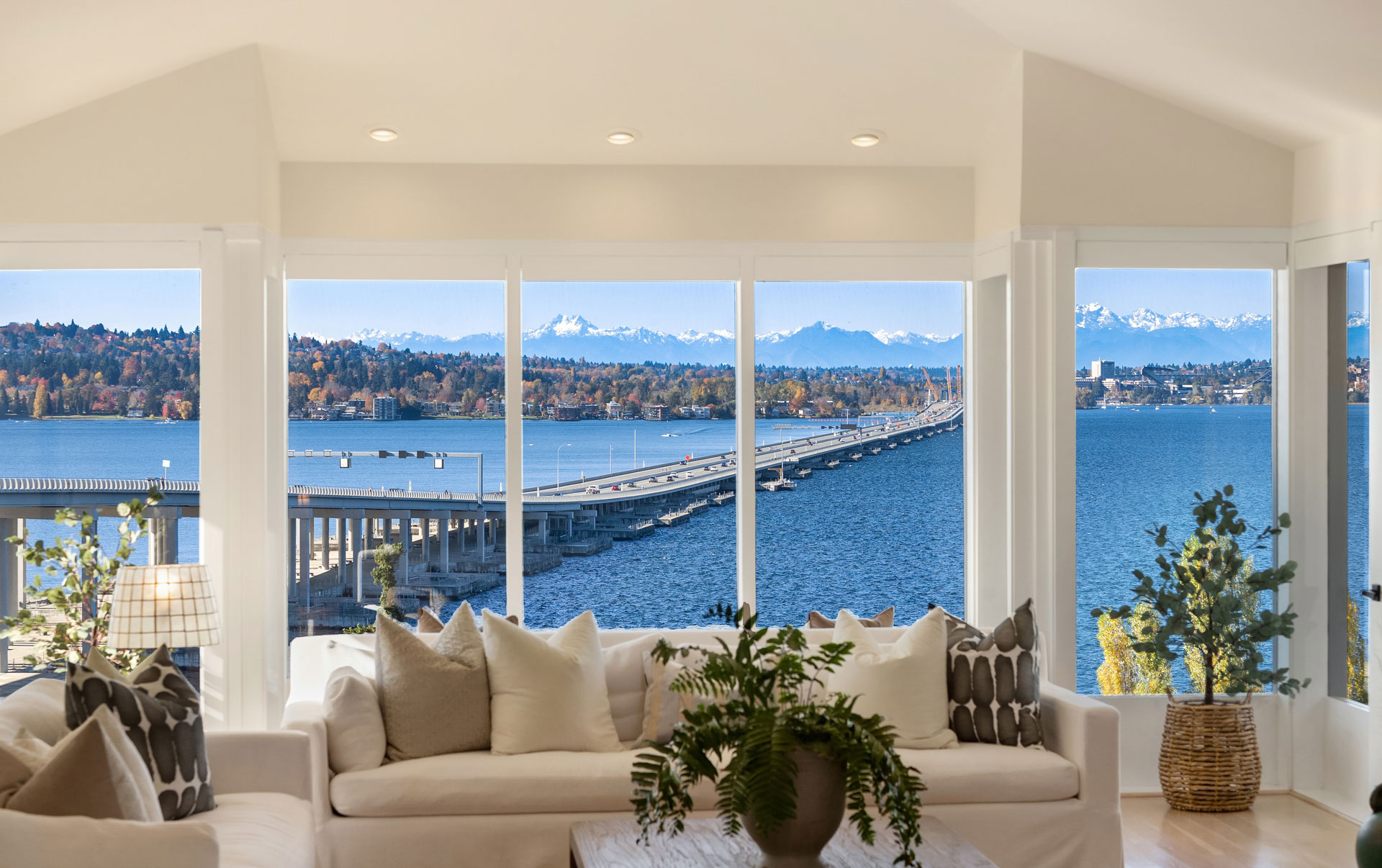 A spacious living room with large windows displaying a stunning view of a lake, bridge, and mountains in the distance. The room features a white sofa with various pillows and indoor plants, creating a cozy and serene atmosphere.